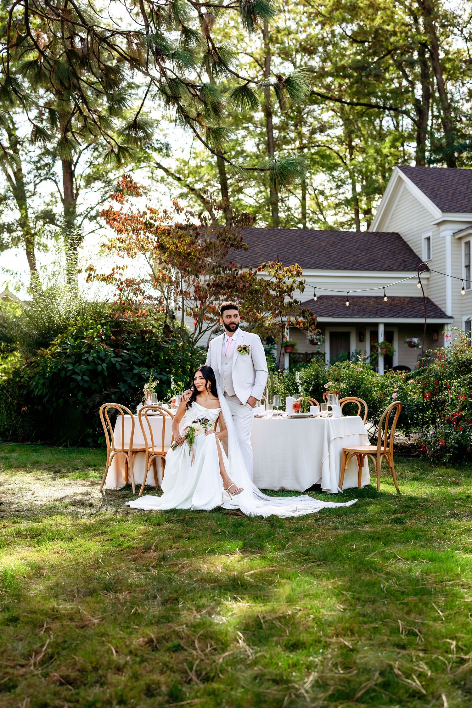 Wedding table setup in a garden with white decor and bistro chairs near a charming house.