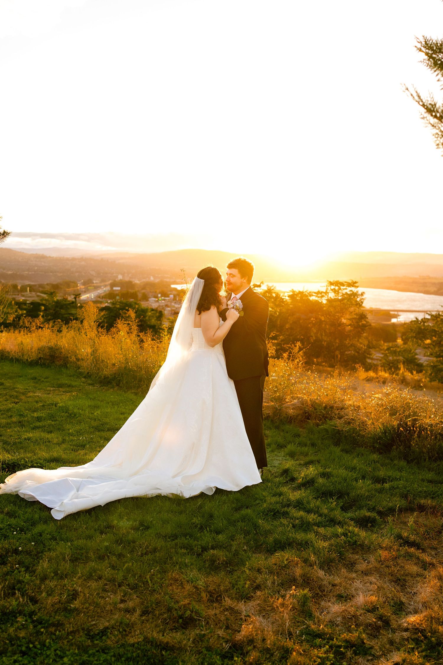 A couple in wedding attire embraces on a hilltop at golden hour with a scenic landscape view in the background.