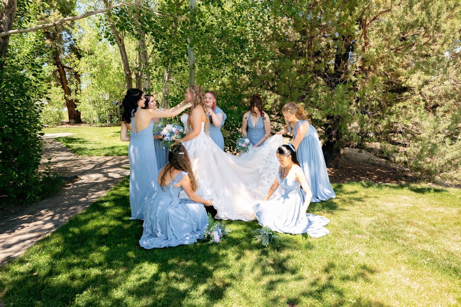 Bridesmaids in light blue dresses help bride with gown on green lawn surrounded by trees.