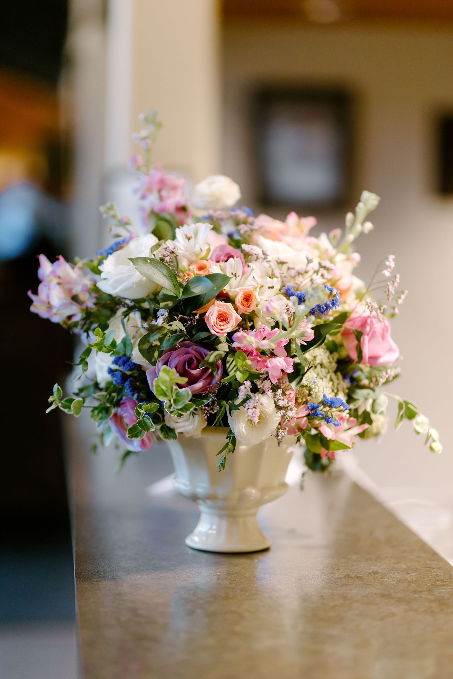 Lush floral arrangement in white urn vase featuring pink roses, purple blooms, and white flowers on a marble surface.