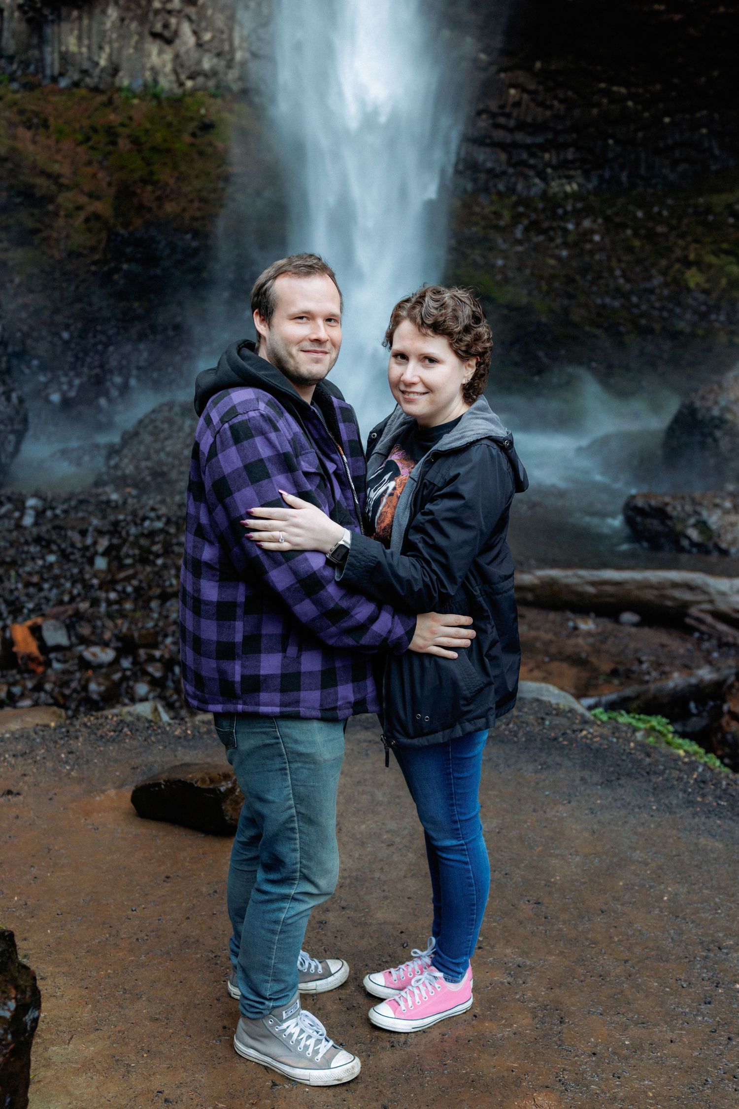 A couple embraces while standing in front of a scenic waterfall on a nature trail.