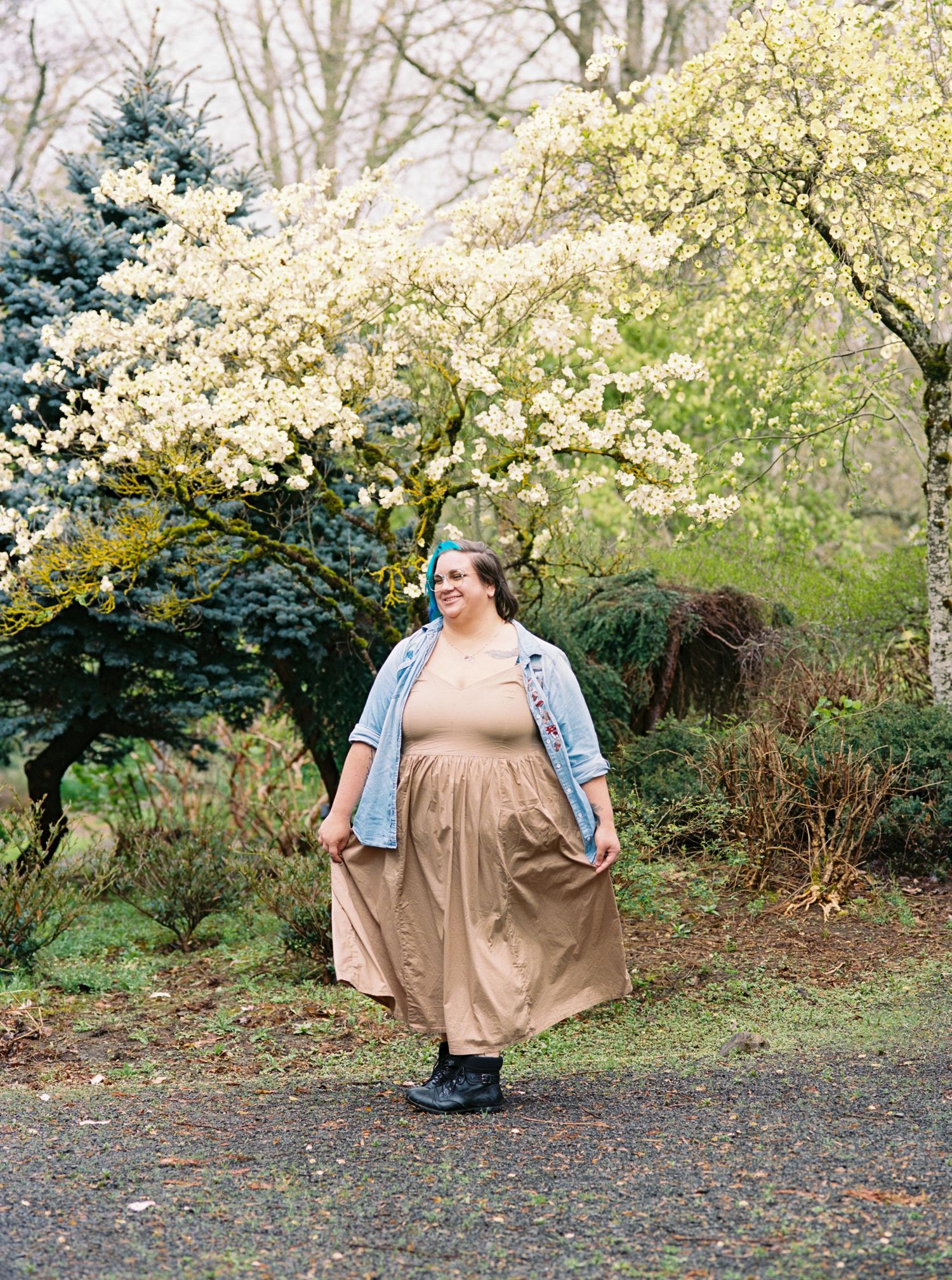 Woman in tan dress and denim jacket walks through a blooming spring garden with white flowering trees.