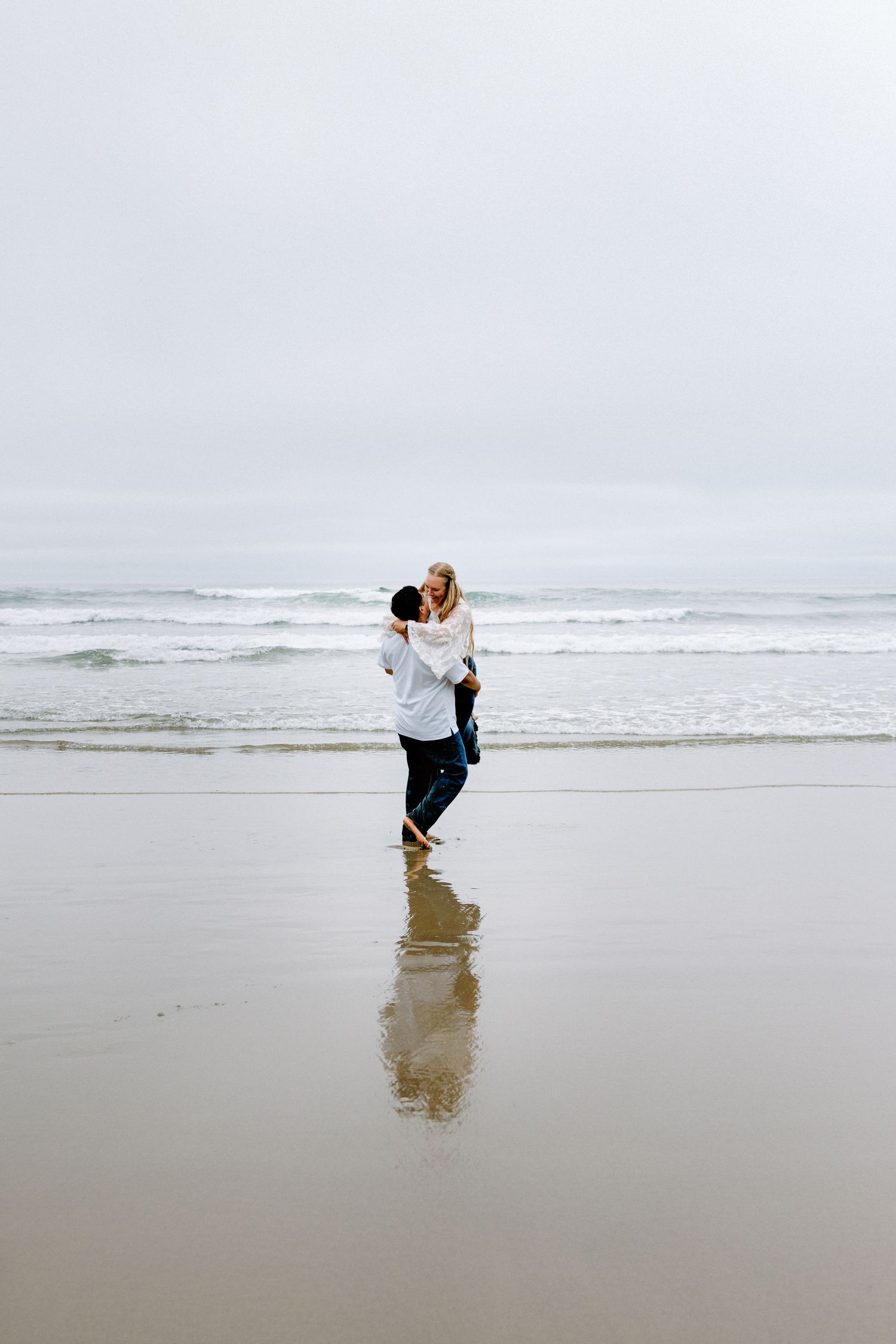 Romantic couple spinning joyfully on wet reflective beach with ocean waves and overcast sky.