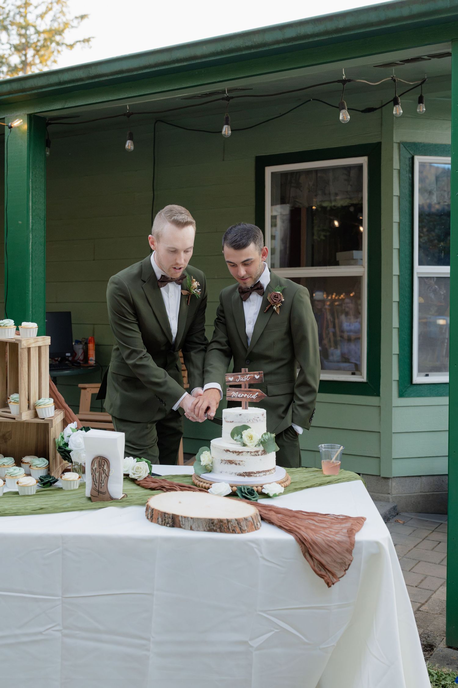 Two grooms in matching green tuxedos cut their rustic naked wedding cake at an outdoor reception.