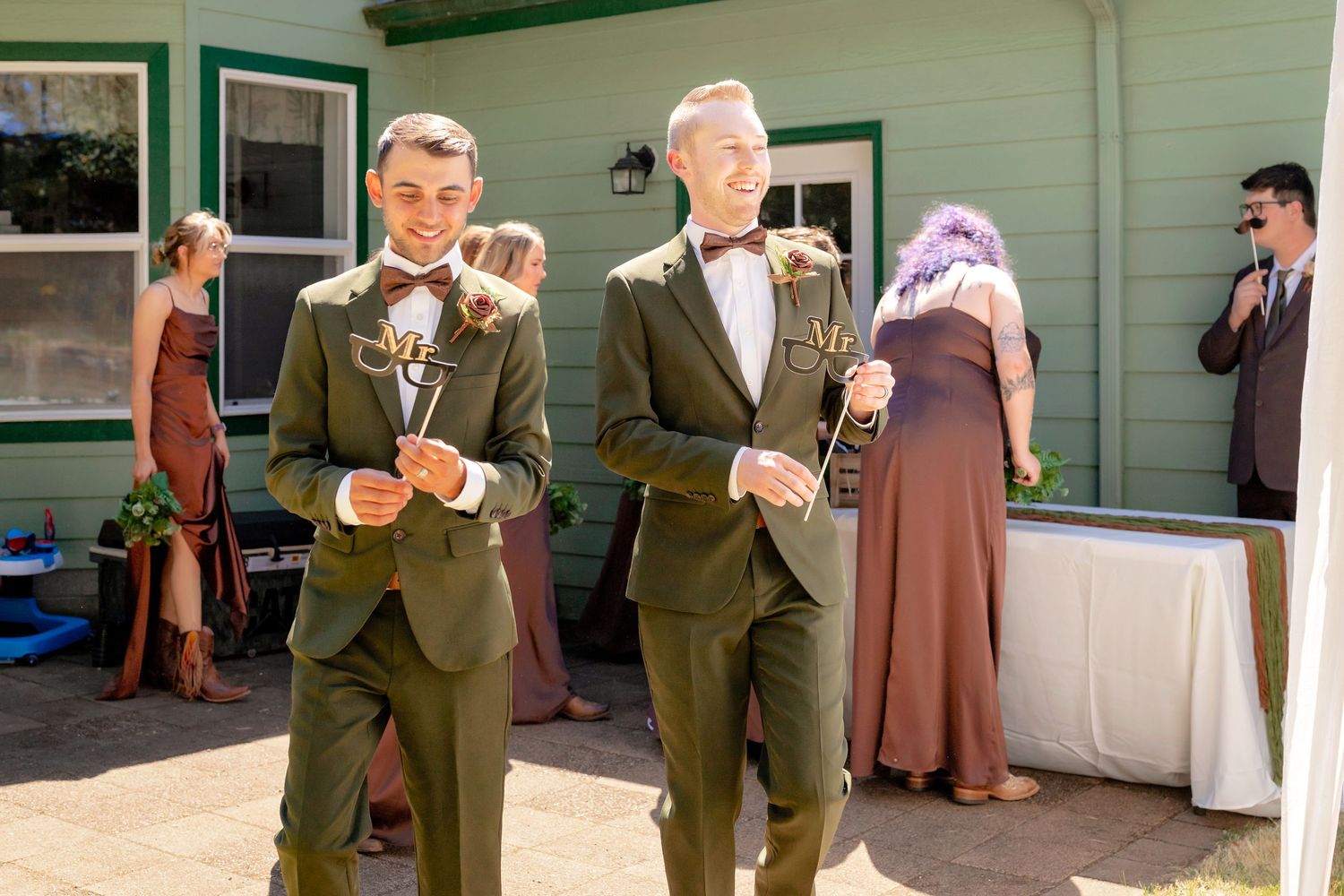 Two grooms in olive green suits with bow ties and boutonnieres smiling at outdoor wedding.