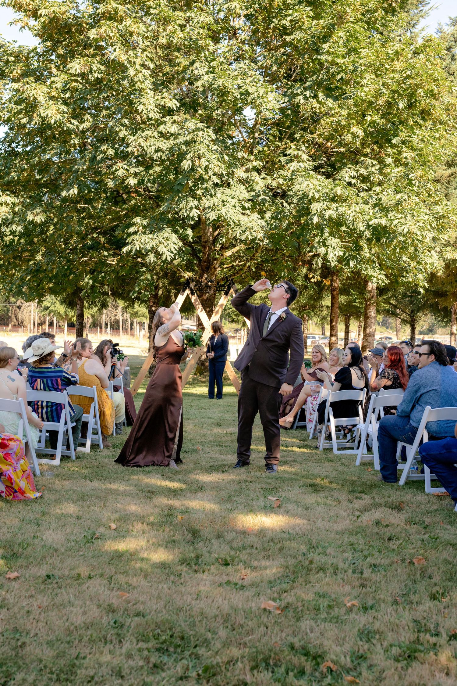 Groom and bridesmaid walk down outdoor wedding aisle amid seated guests and lush green trees.