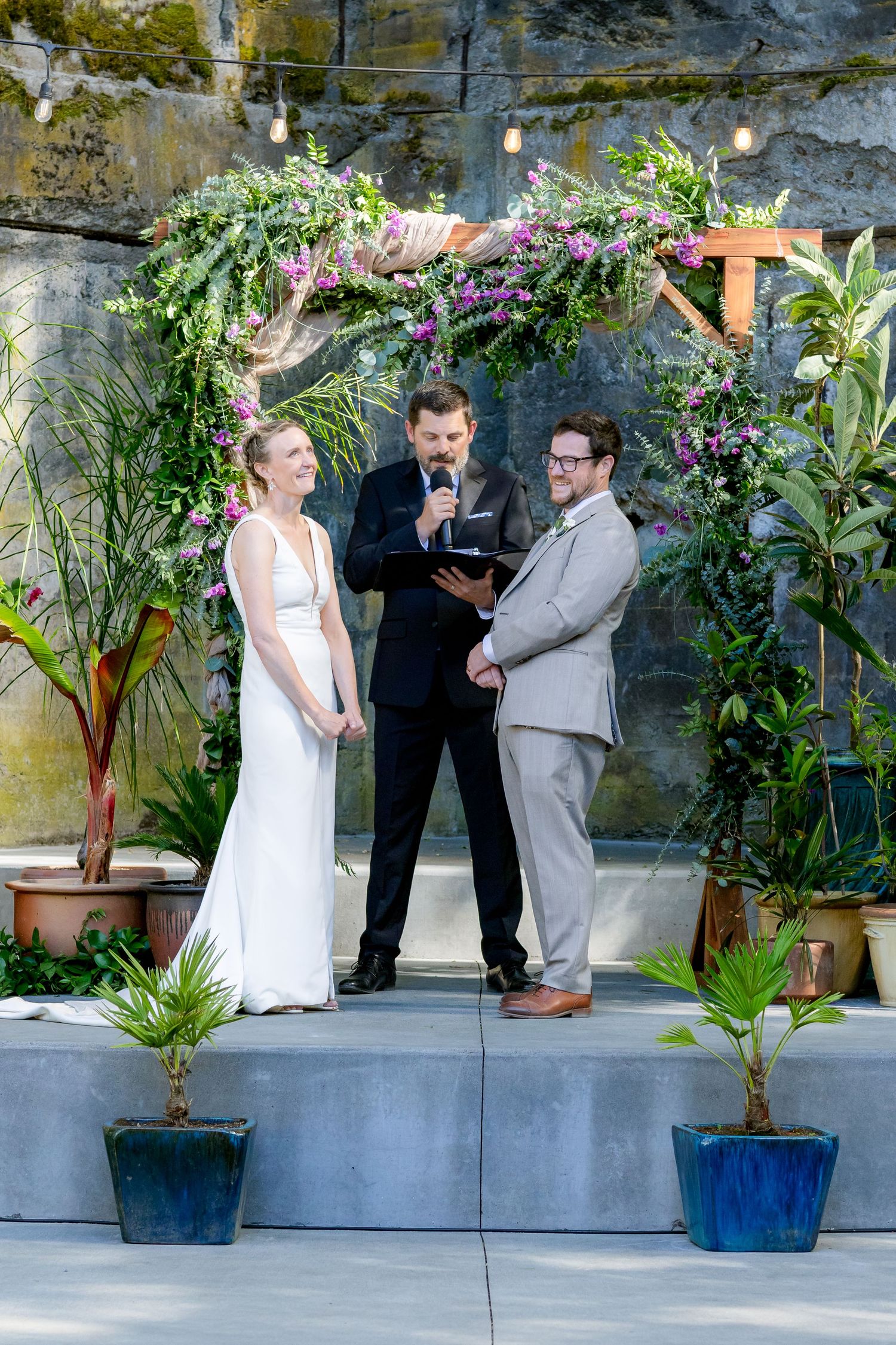 Bride and groom exchange vows under floral arch with officiant at garden wedding ceremony.