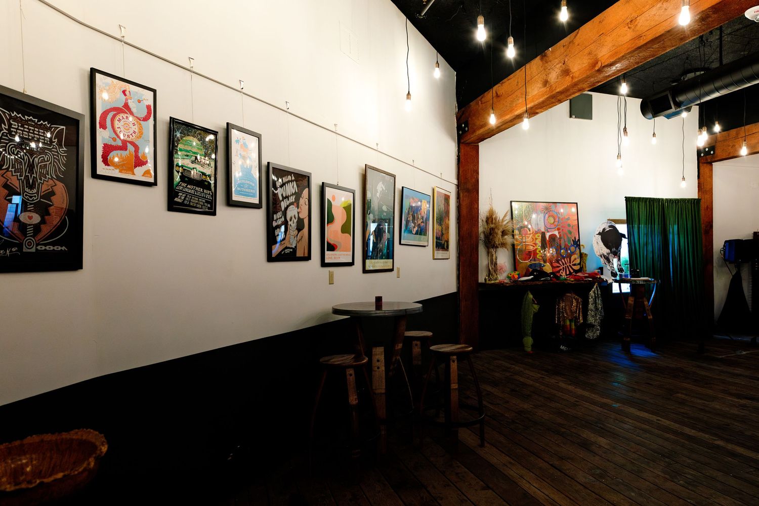 Art gallery interior with framed posters on white walls, wooden stools, string lights, and visitors browsing artwork.