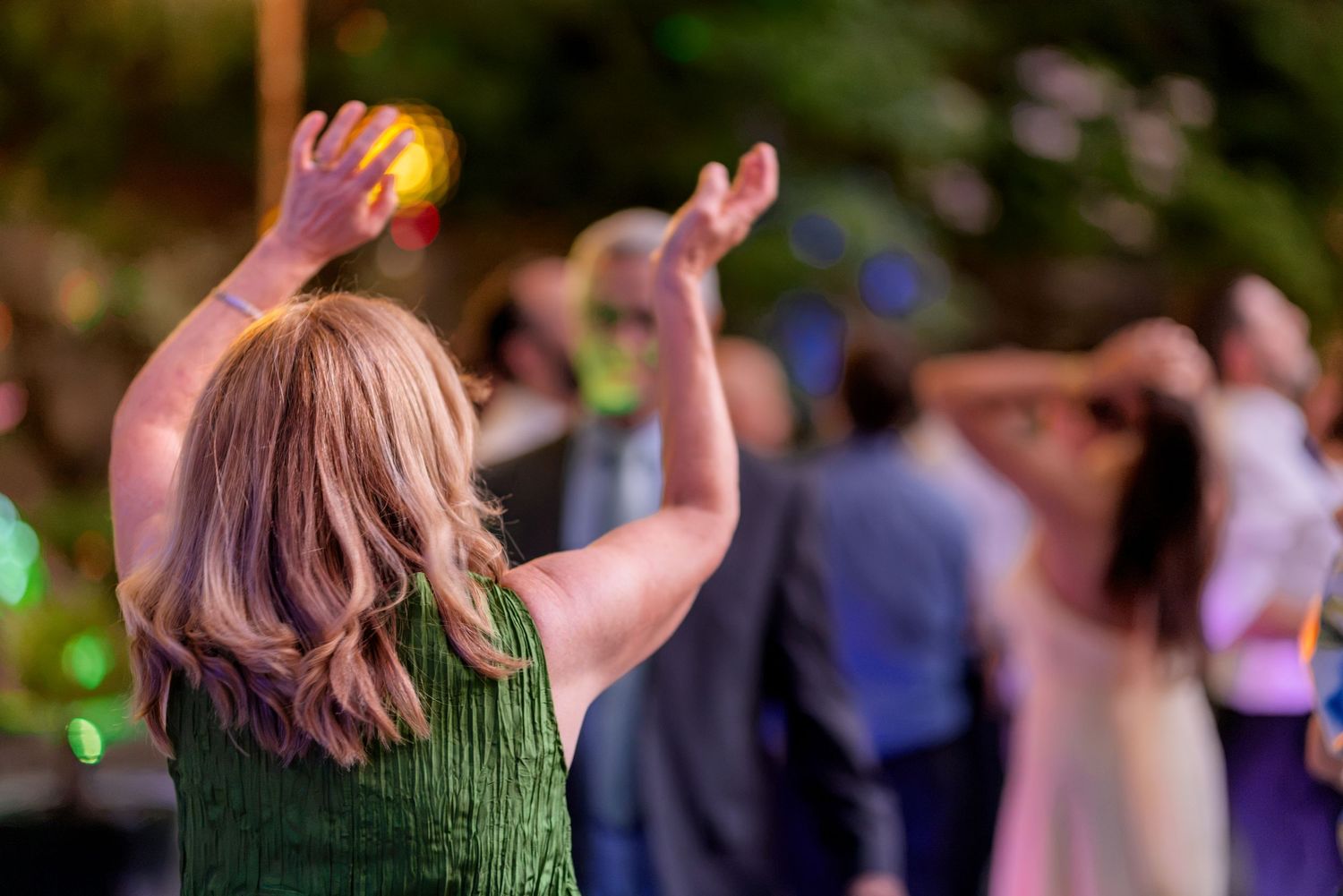 Wedding guest in green dress raises arms while dancing at an outdoor evening reception.