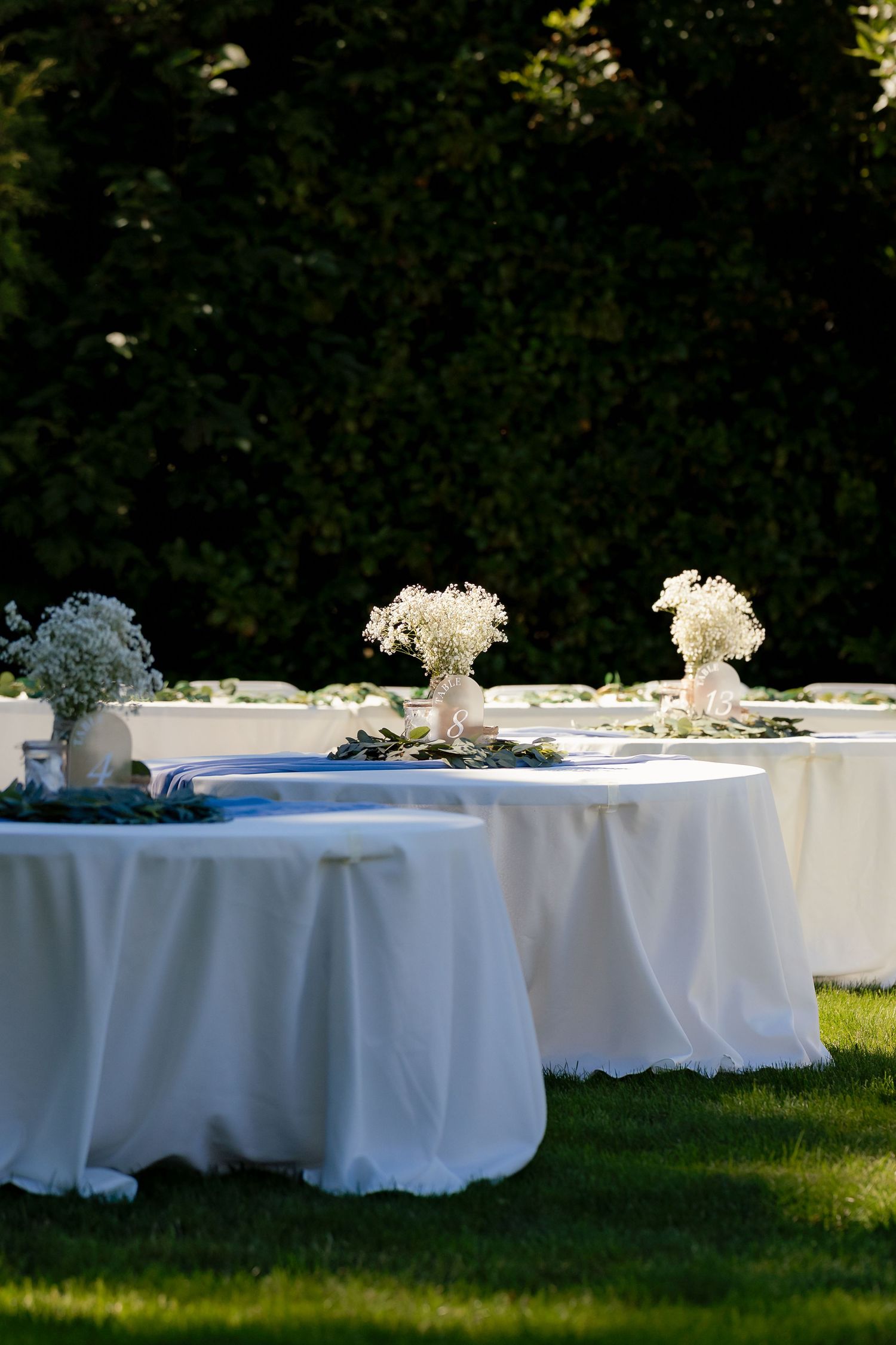 Outdoor wedding reception tables with white linen tablecloths and baby's breath centerpieces on green lawn.
