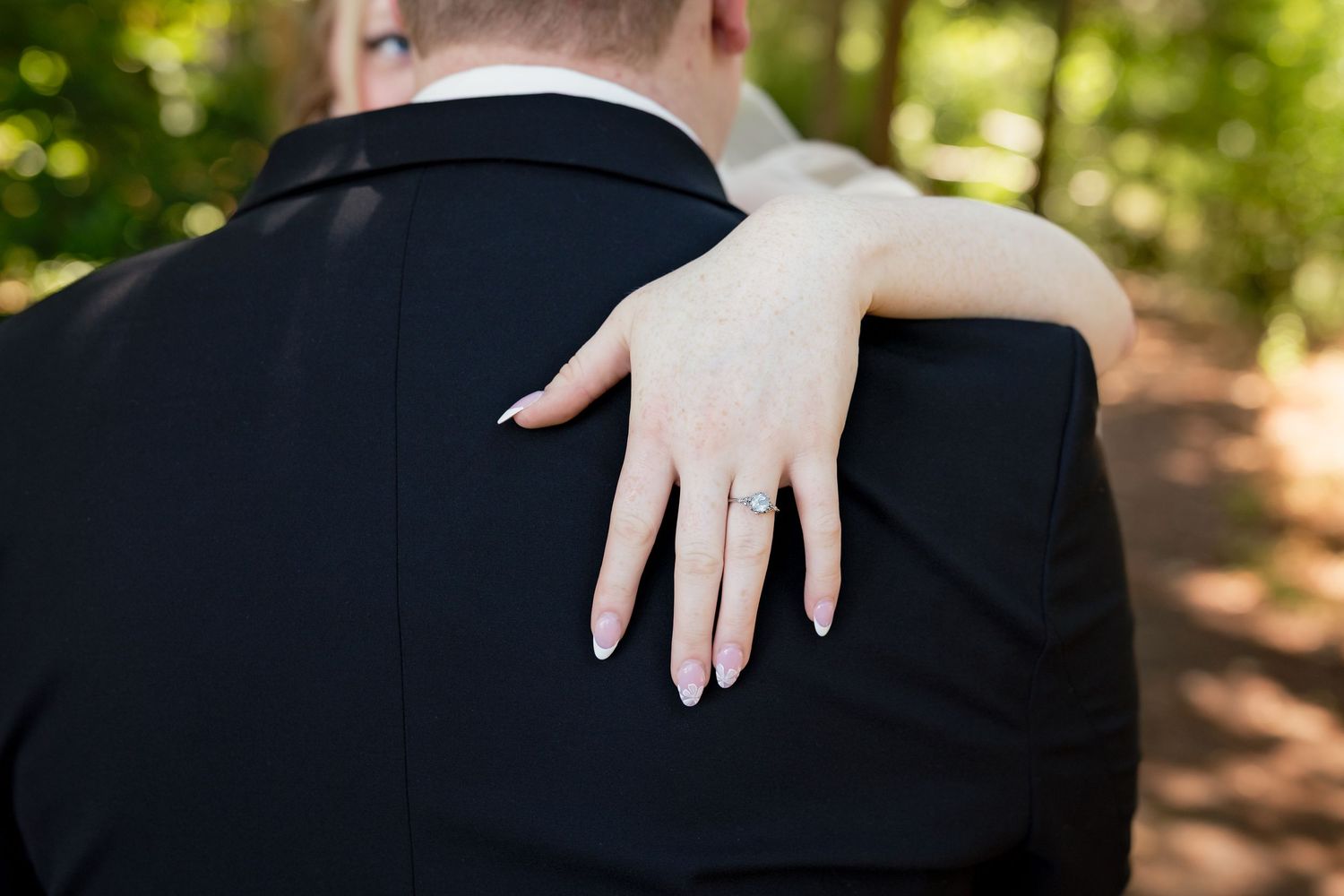 Bride resting her hand on groom's shoulder, showcasing diamond engagement ring, with lush green trees in background.