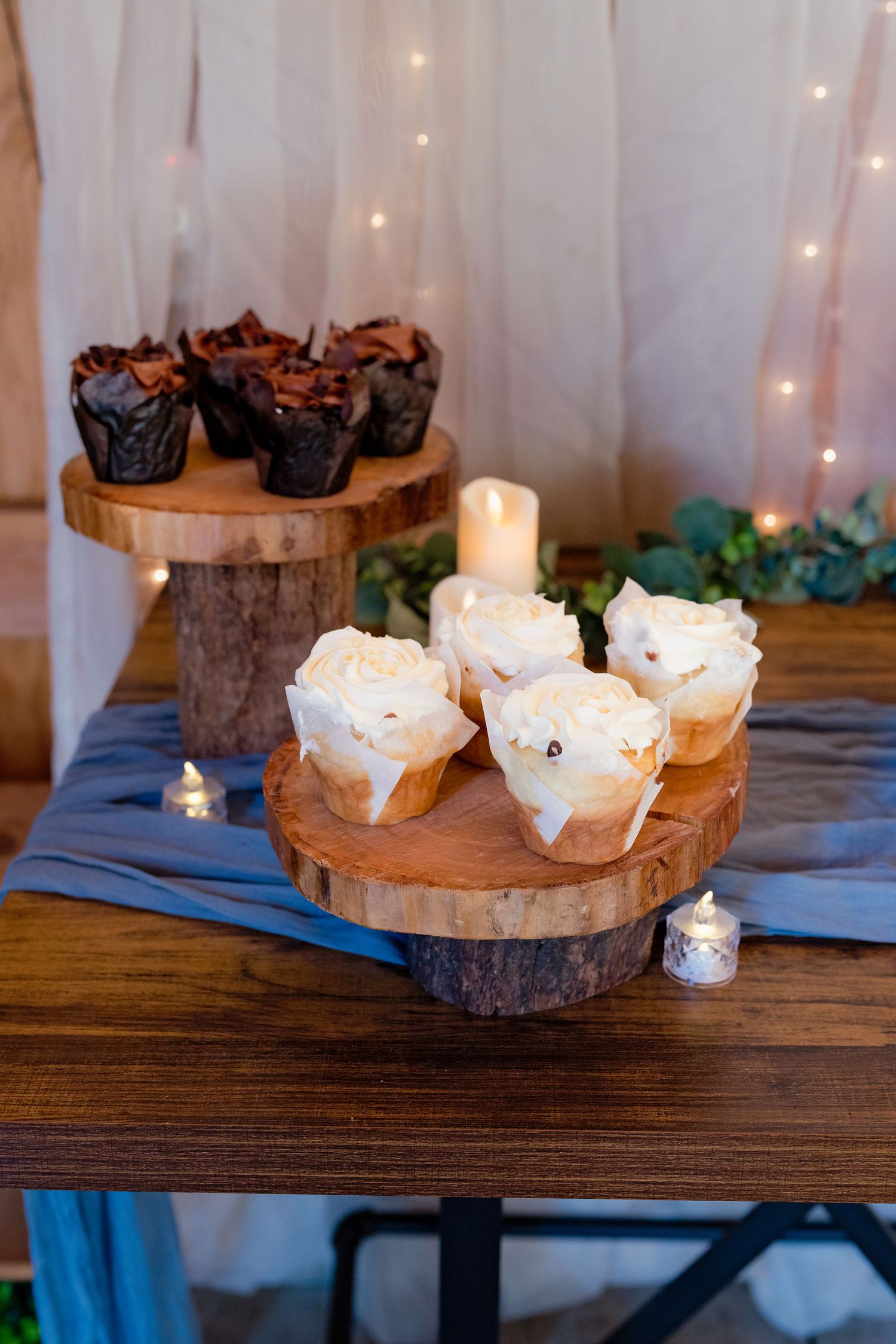 Rustic wedding dessert display with vanilla and chocolate cupcakes on wooden cake stands with candles.