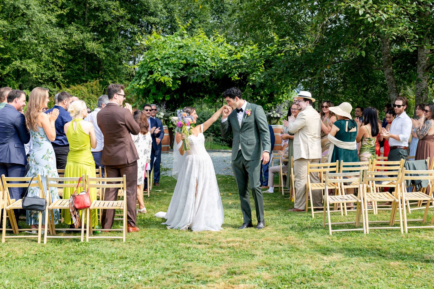 Newlywed couple sharing a kiss at an outdoor garden wedding ceremony with guests applauding on a sunny day.