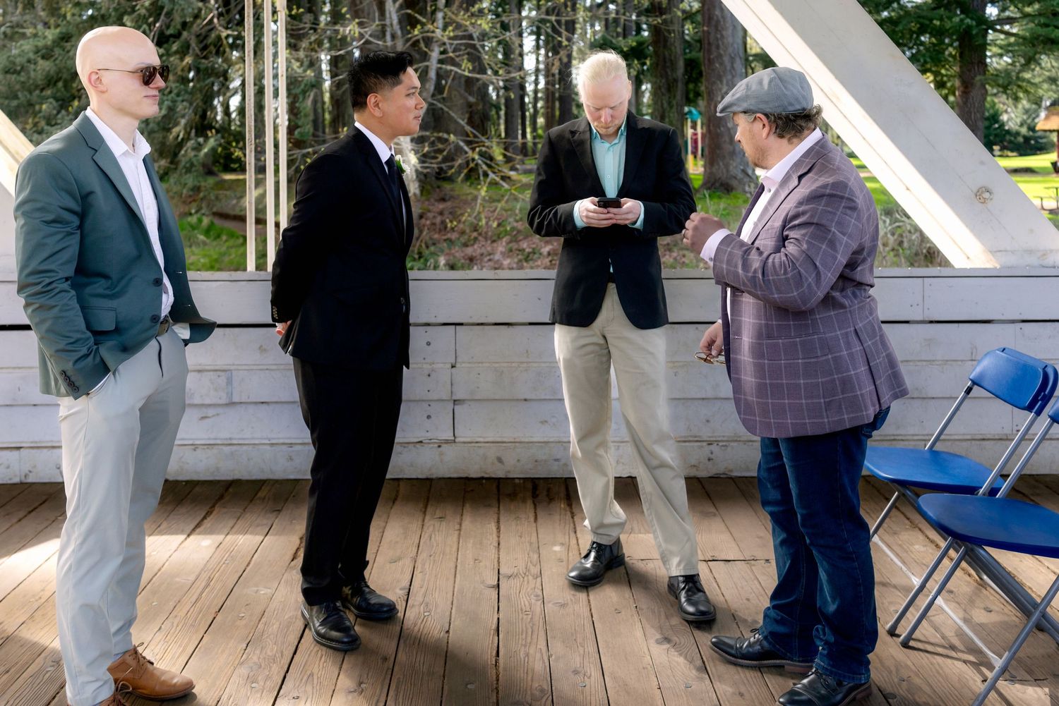 Four men in business casual attire stand conversing on an outdoor wooden deck surrounded by trees.