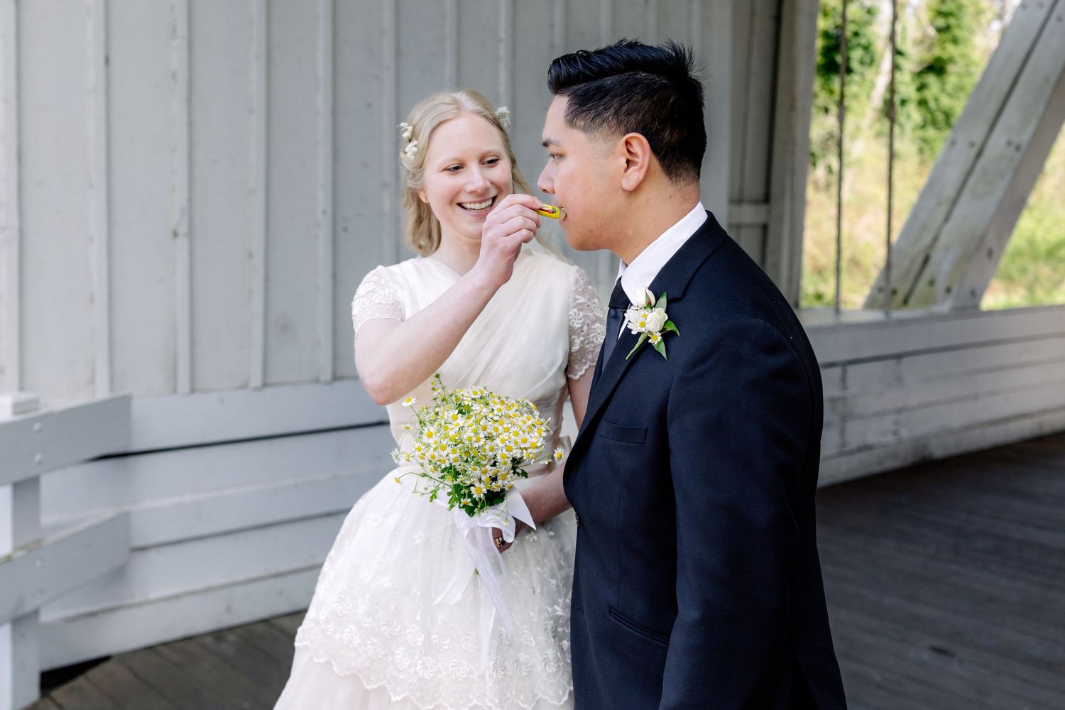 Bride in white lace dress playfully feeds groom a flower petal at outdoor wedding venue.
