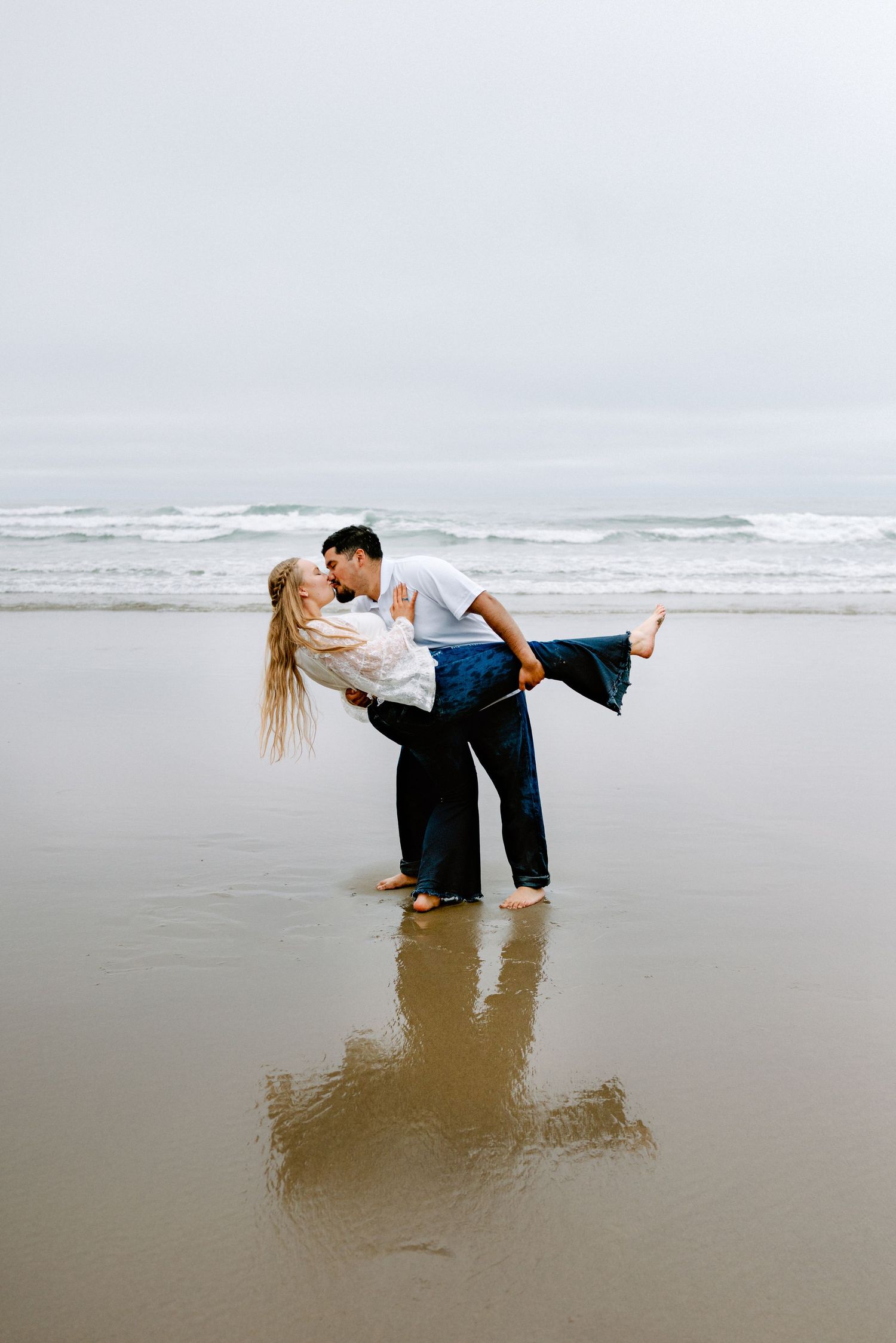 Man dipping woman for a kiss on wet reflective sand with ocean waves behind them.