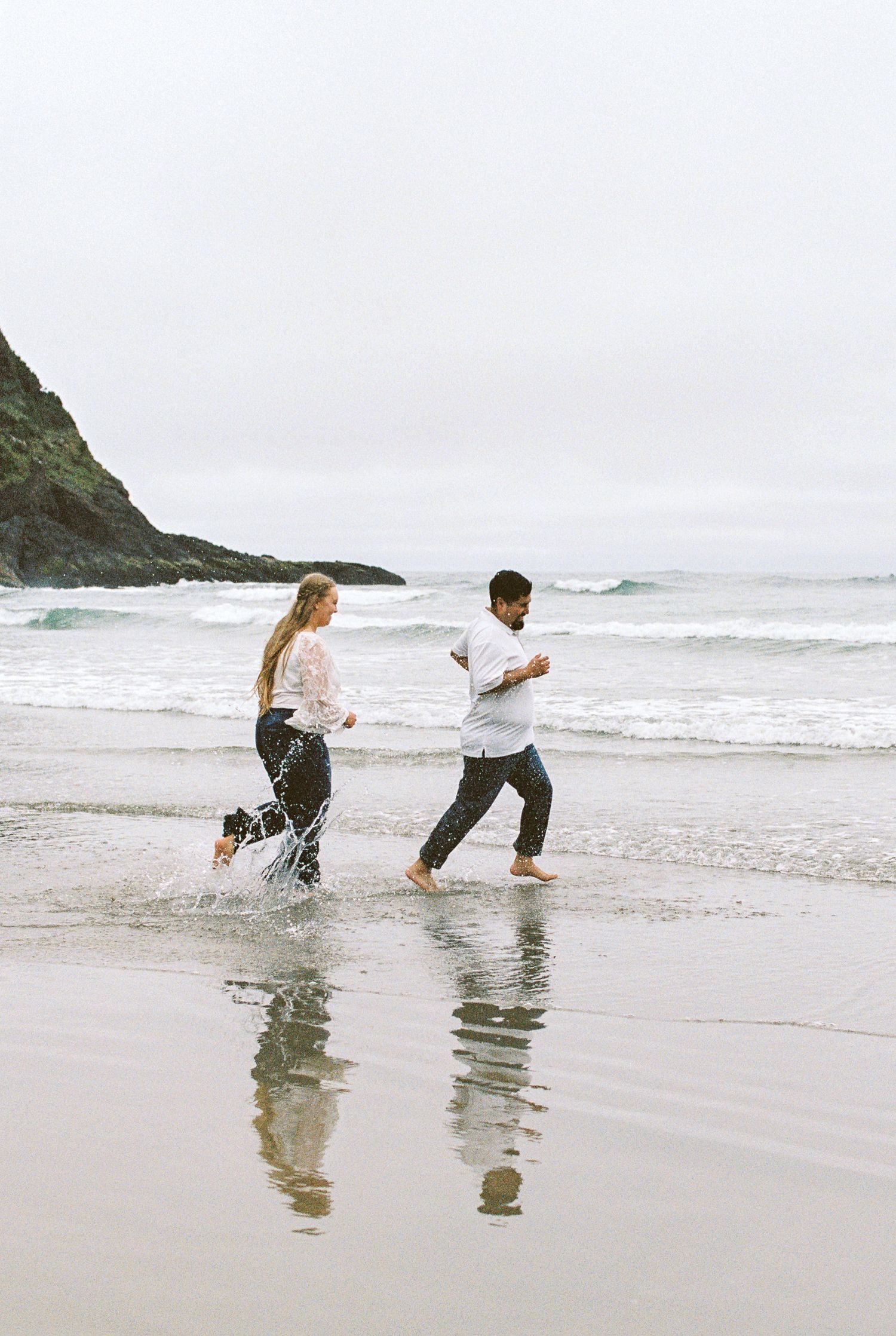 Joyful couple running barefoot through ocean waves on a scenic beach with rocky headlands.