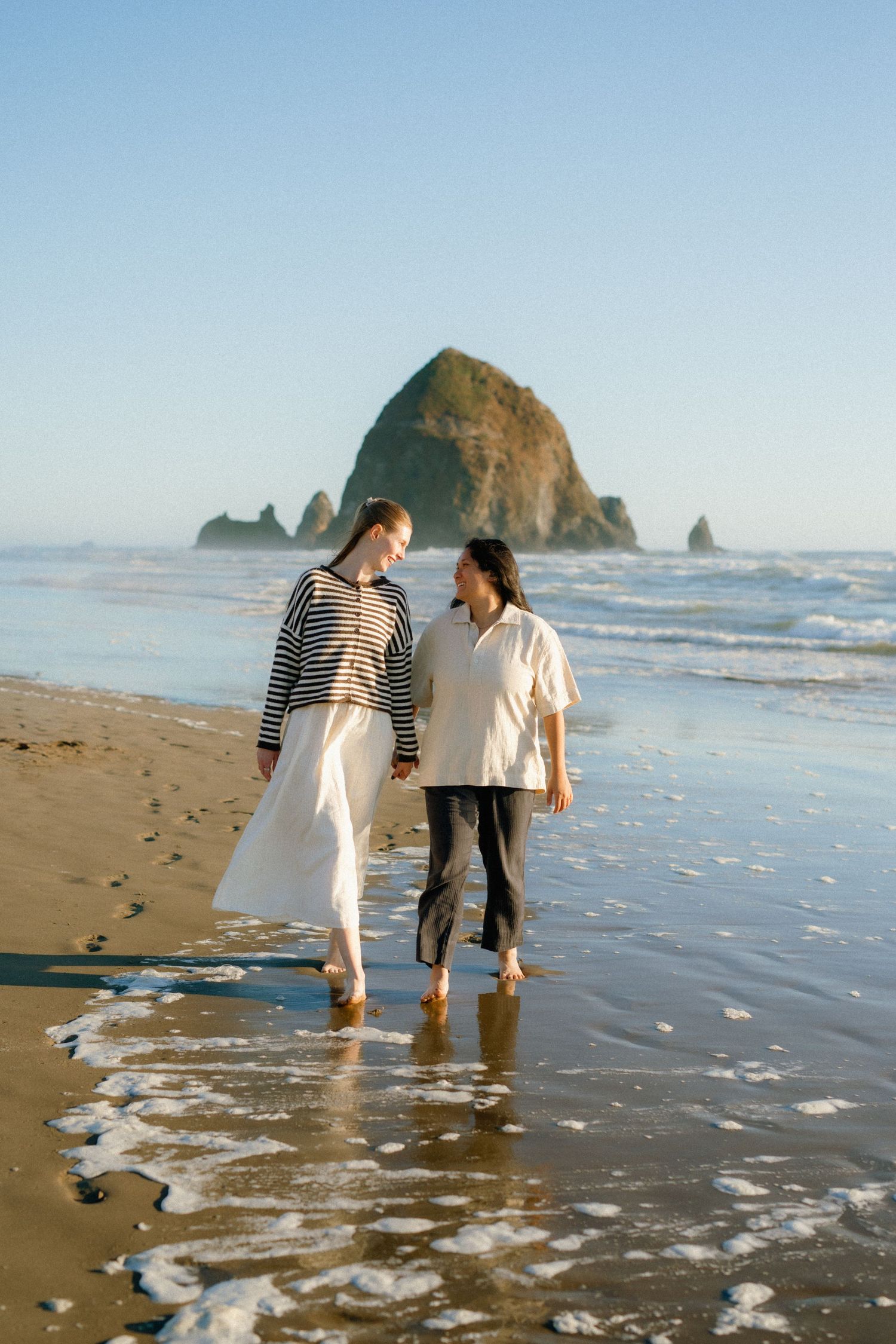 Two women walk hand-in-hand along a sandy beach with Haystack Rock in the background at Cannon Beach, Oregon.