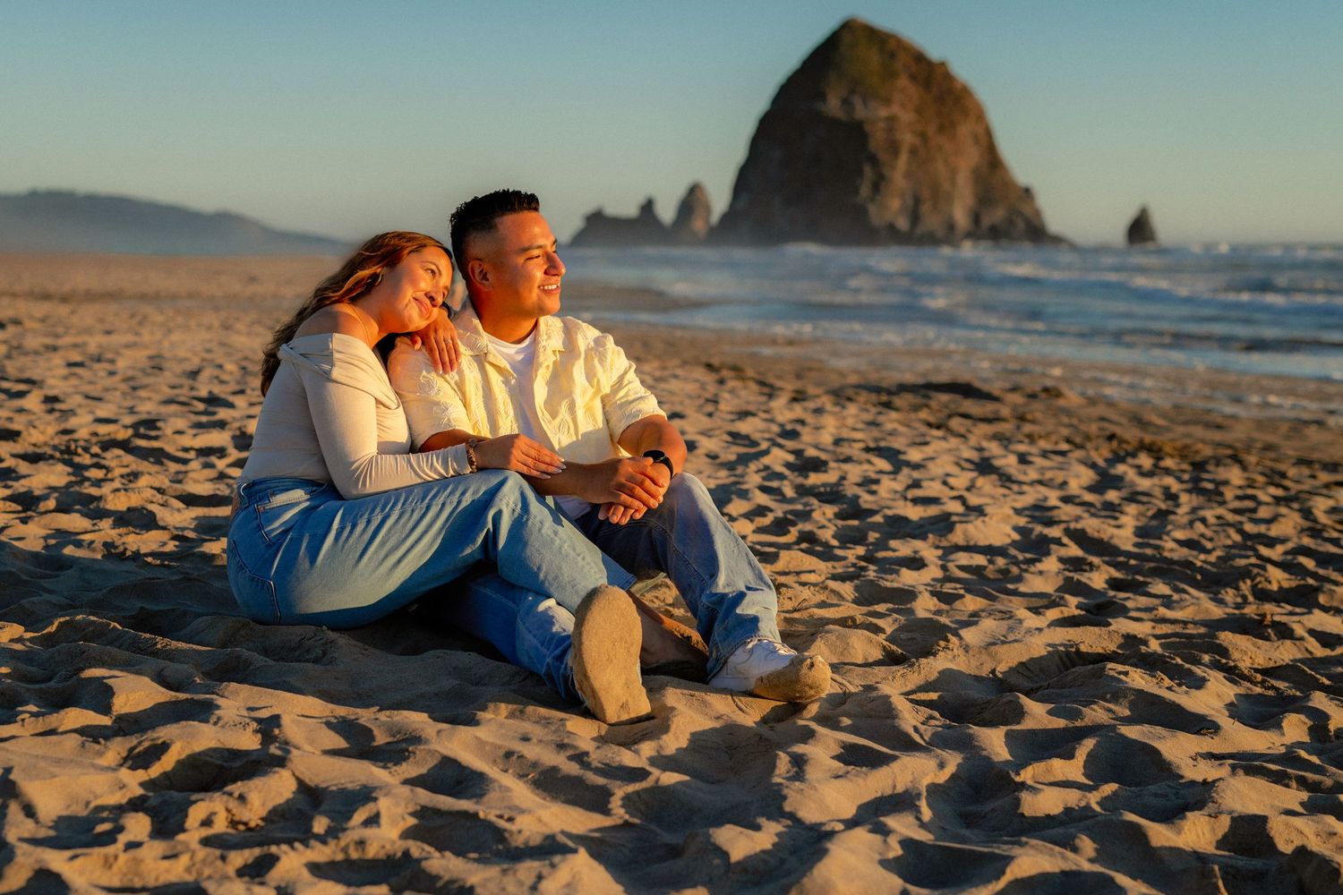 Couple sitting together on sandy beach at sunset with iconic Haystack Rock in background at Cannon Beach Oregon.