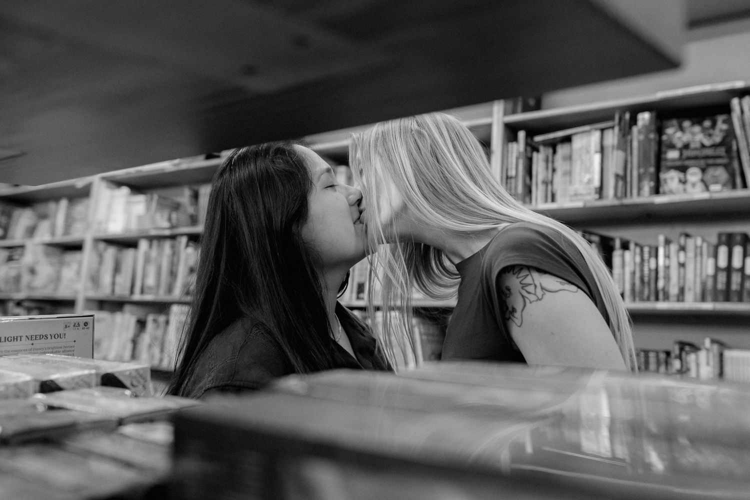 Two women sharing a kiss in a bookstore, captured in black and white photography.