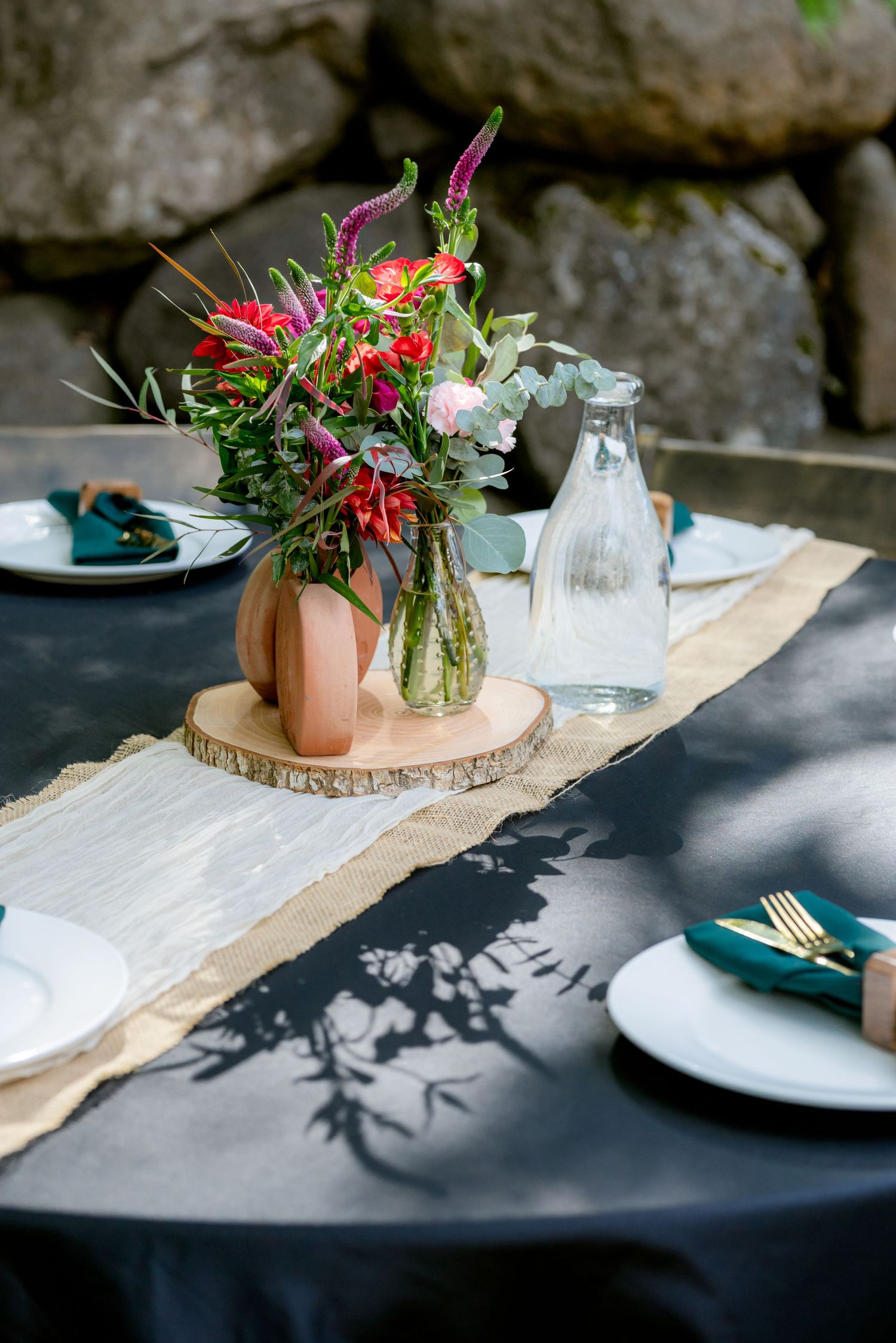 Outdoor dining table with floral centerpiece, burlap runner, white plates, gold cutlery, and teal napkins against stone wall.