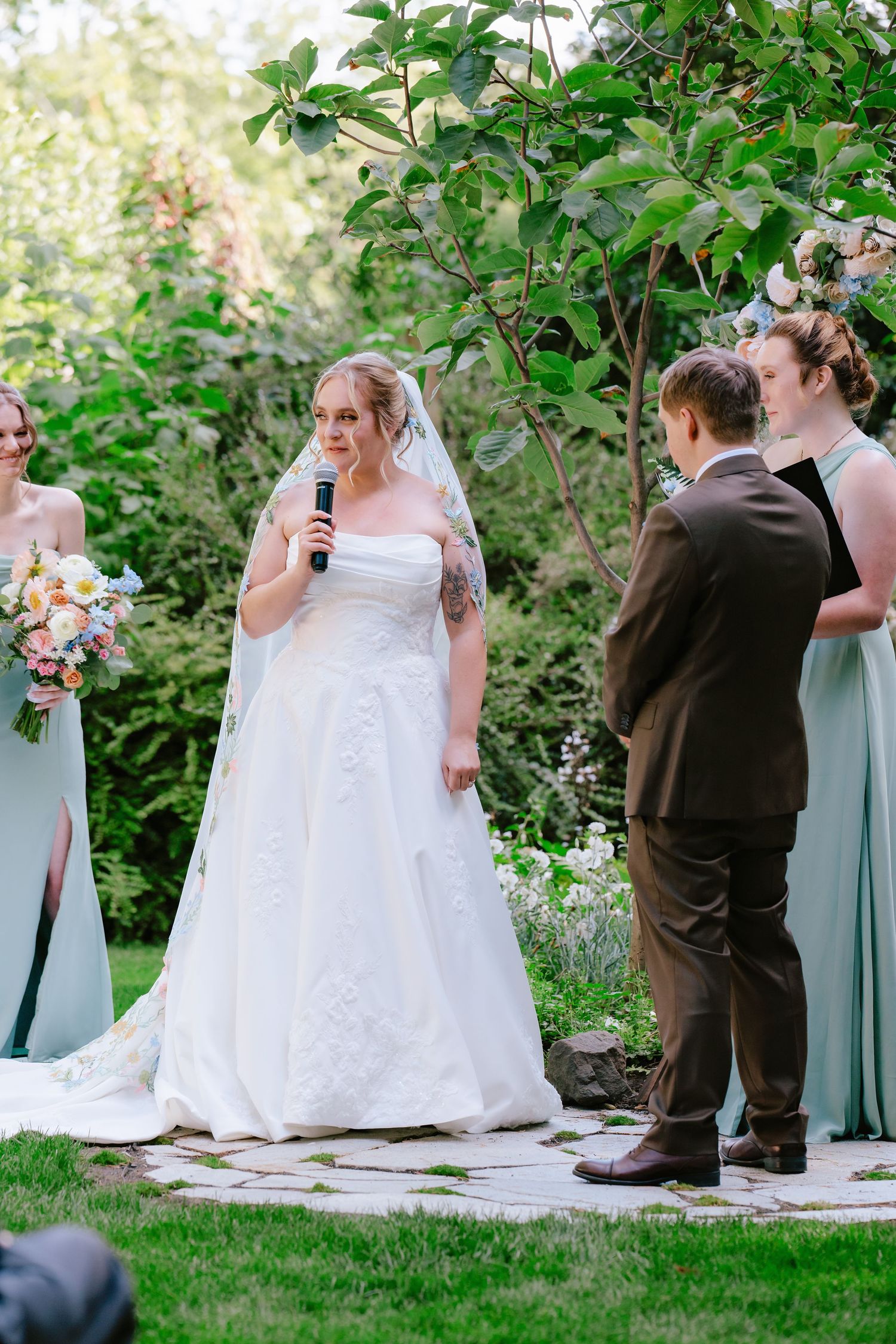 Bride in white strapless gown recites vows holding microphone at outdoor garden wedding ceremony.