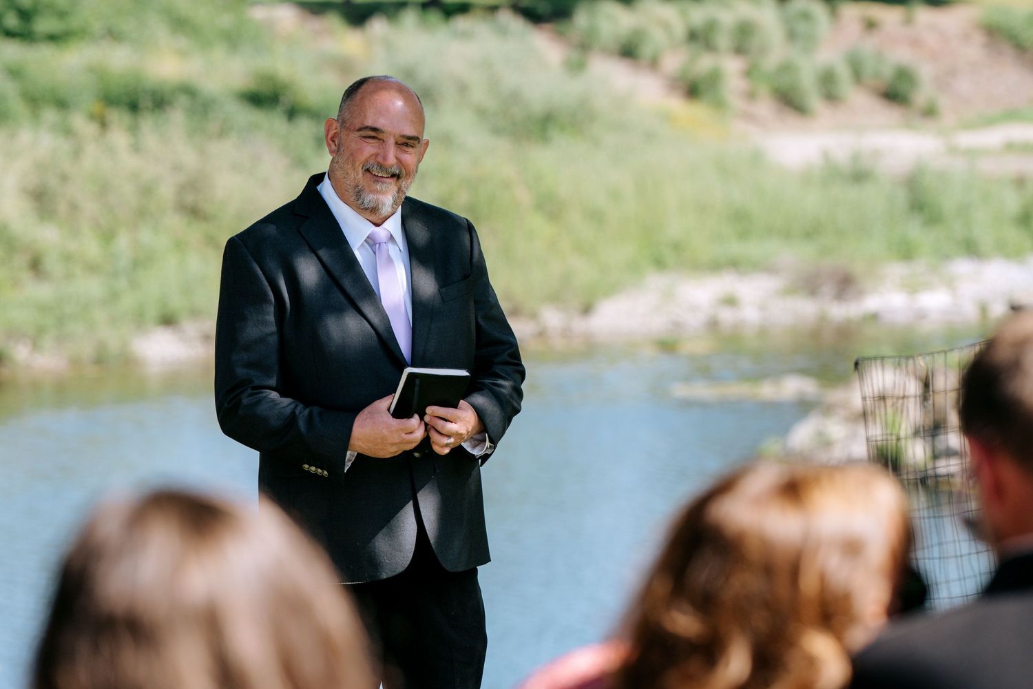 Smiling wedding officiant in a black suit holds a book while addressing guests at an outdoor waterside ceremony.