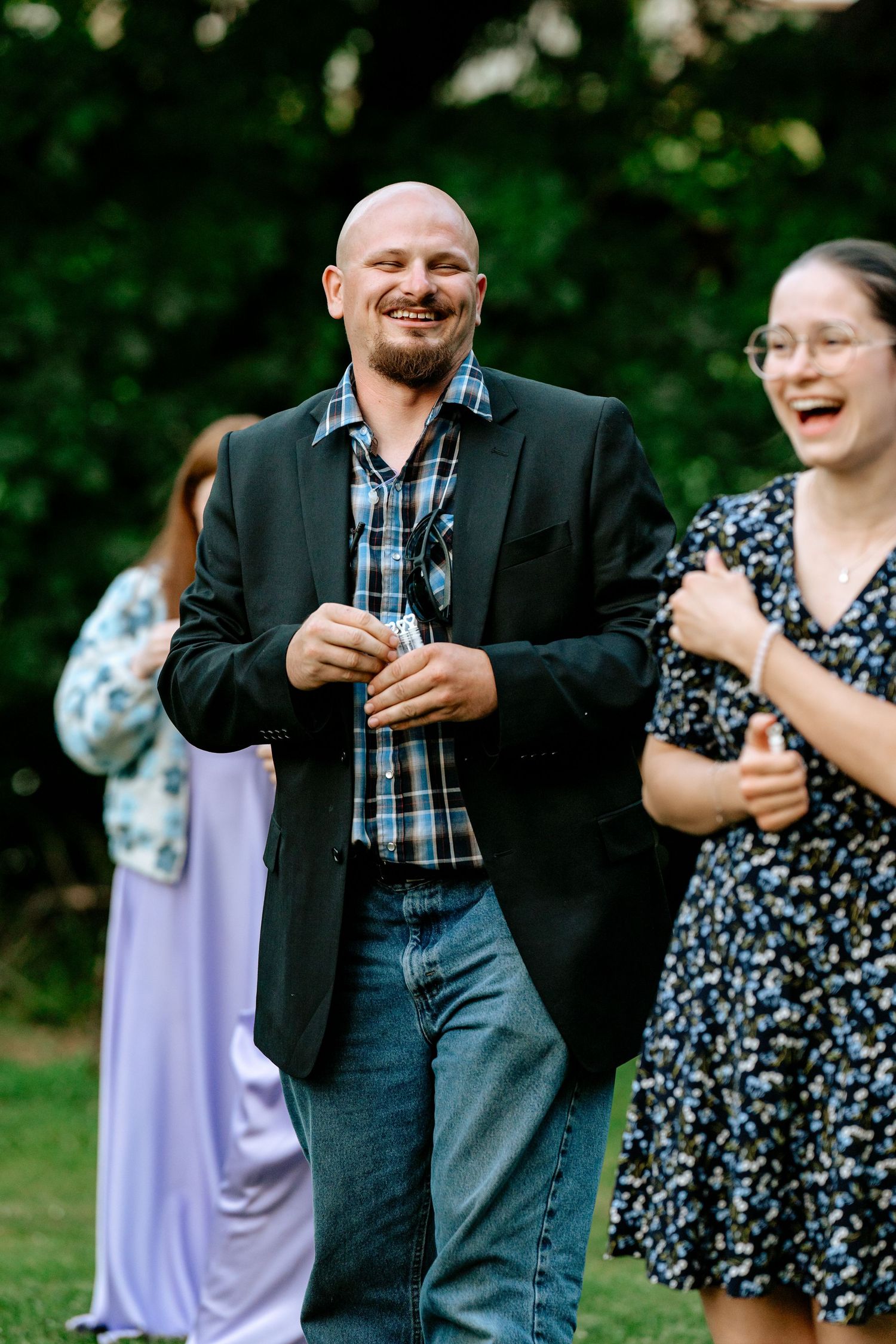 Smiling bald man in black blazer and plaid shirt laughs with guests at an outdoor wedding reception.