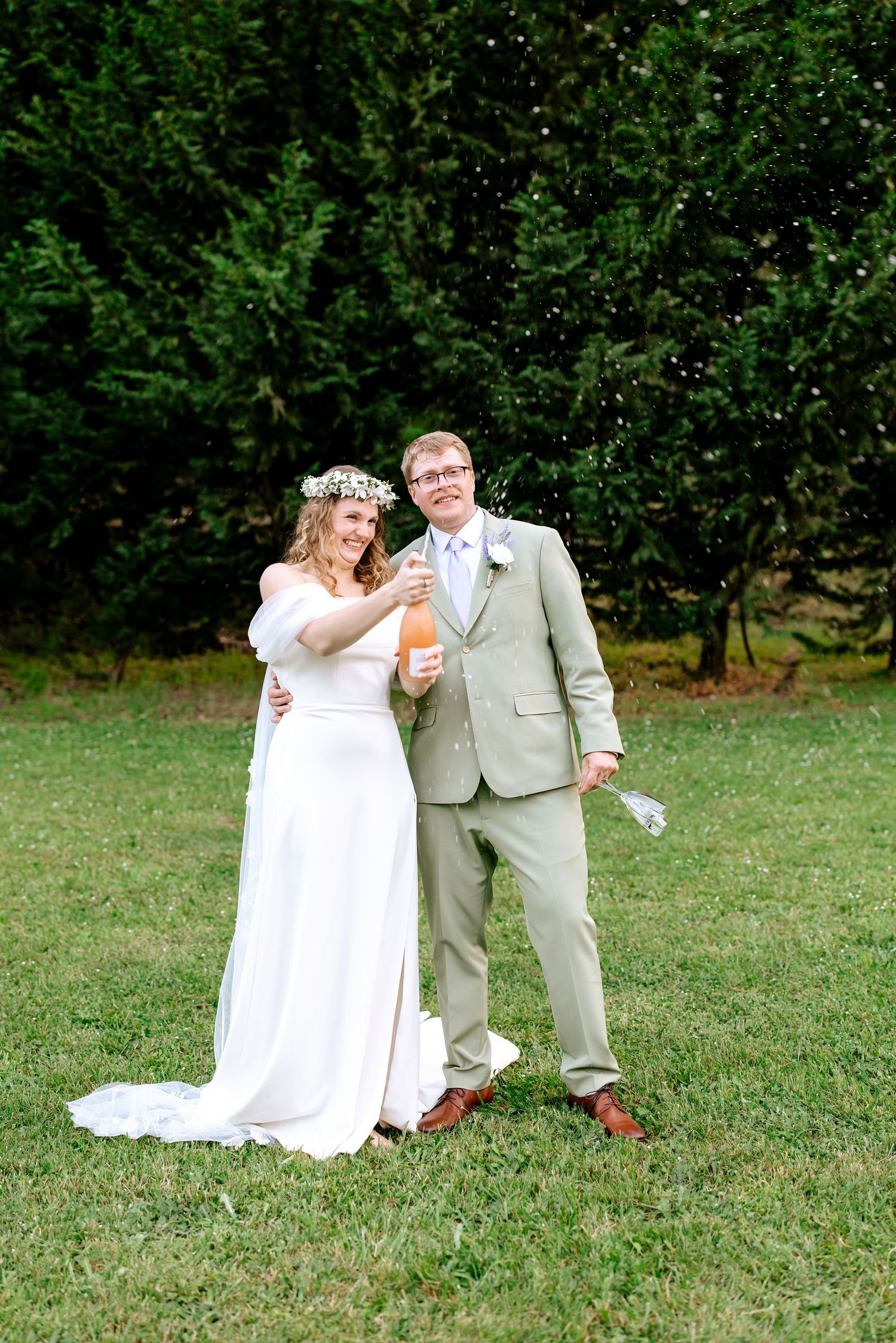 Smiling bride and groom pop champagne bottle together on green lawn after outdoor wedding ceremony.