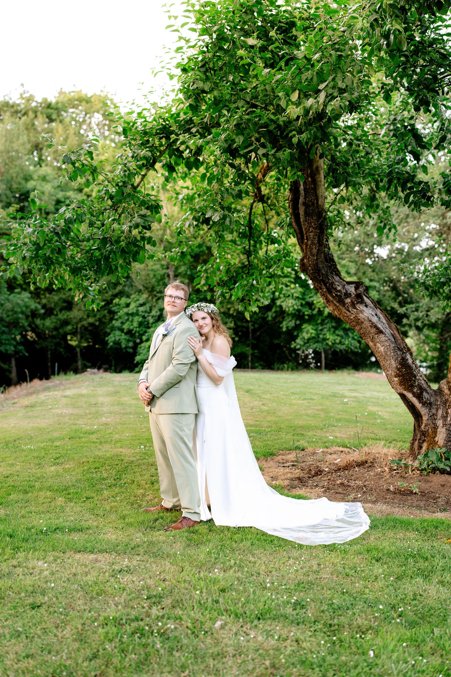 Bride and groom pose elegantly beneath a large leafy tree in a lush green garden.
