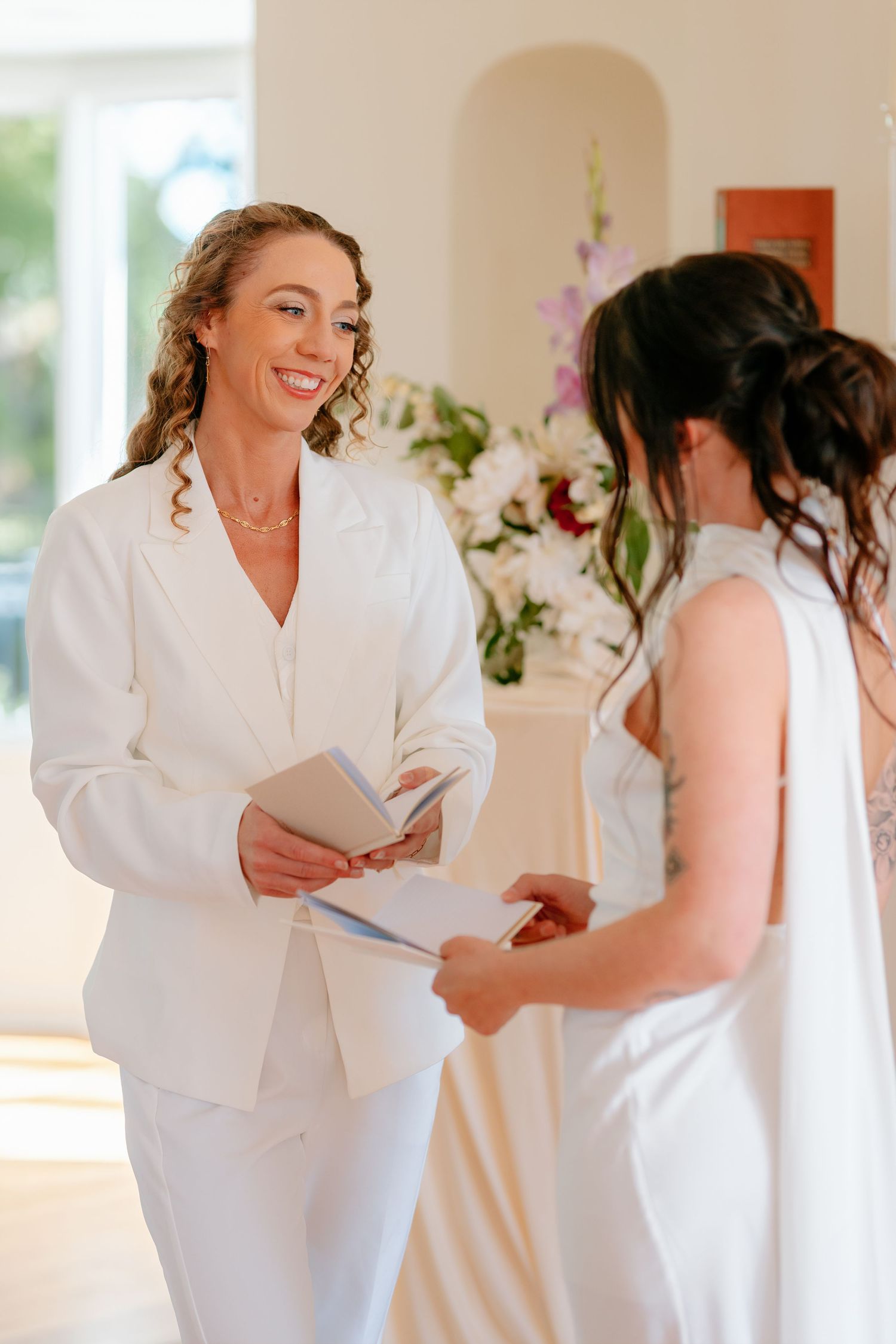 Two brides in white exchange vows indoors, one smiling warmly while holding wedding ceremony booklets near floral arrangements.