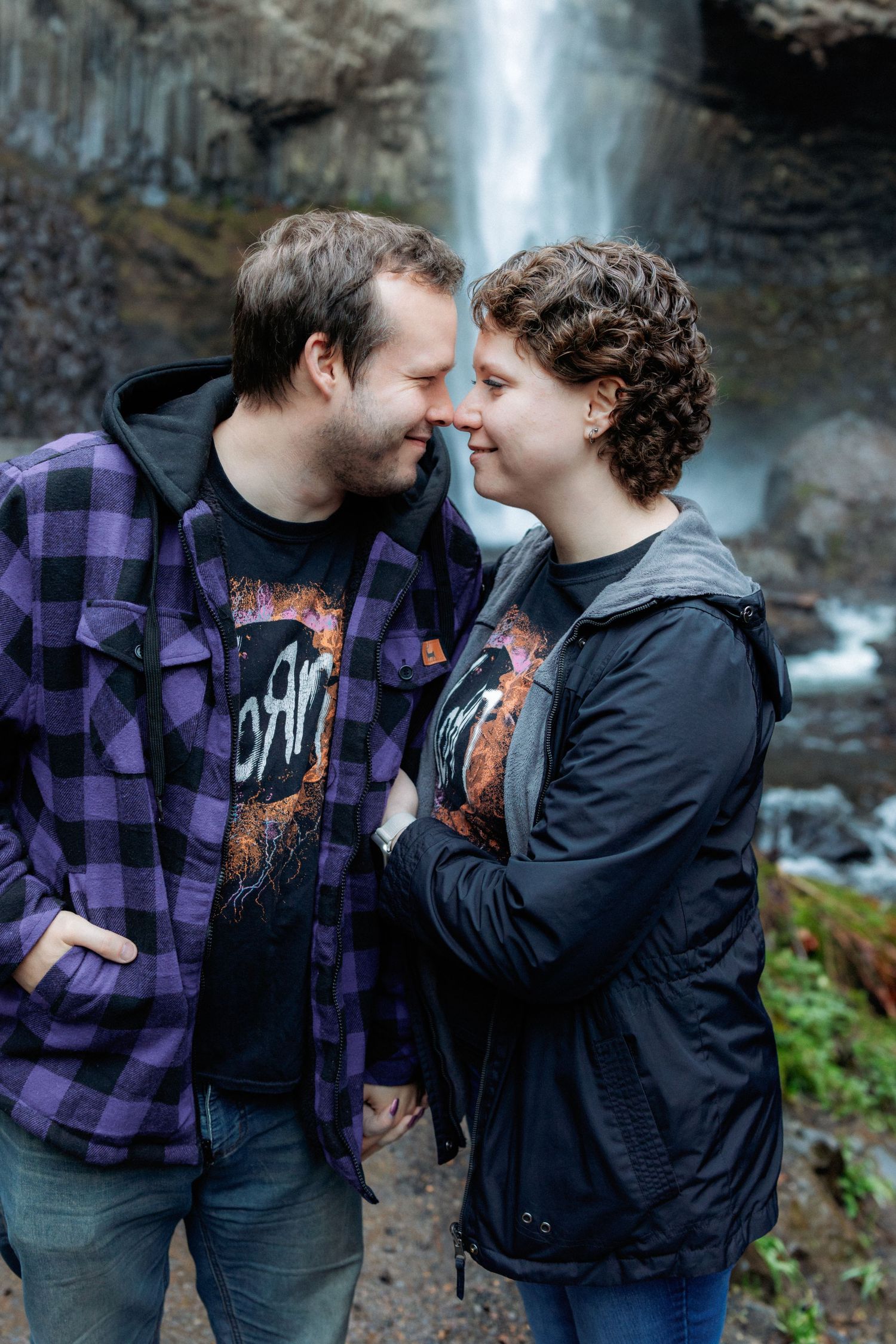 A couple shares intimate moments in front of a waterfall while wearing casual plaid and dark clothing.