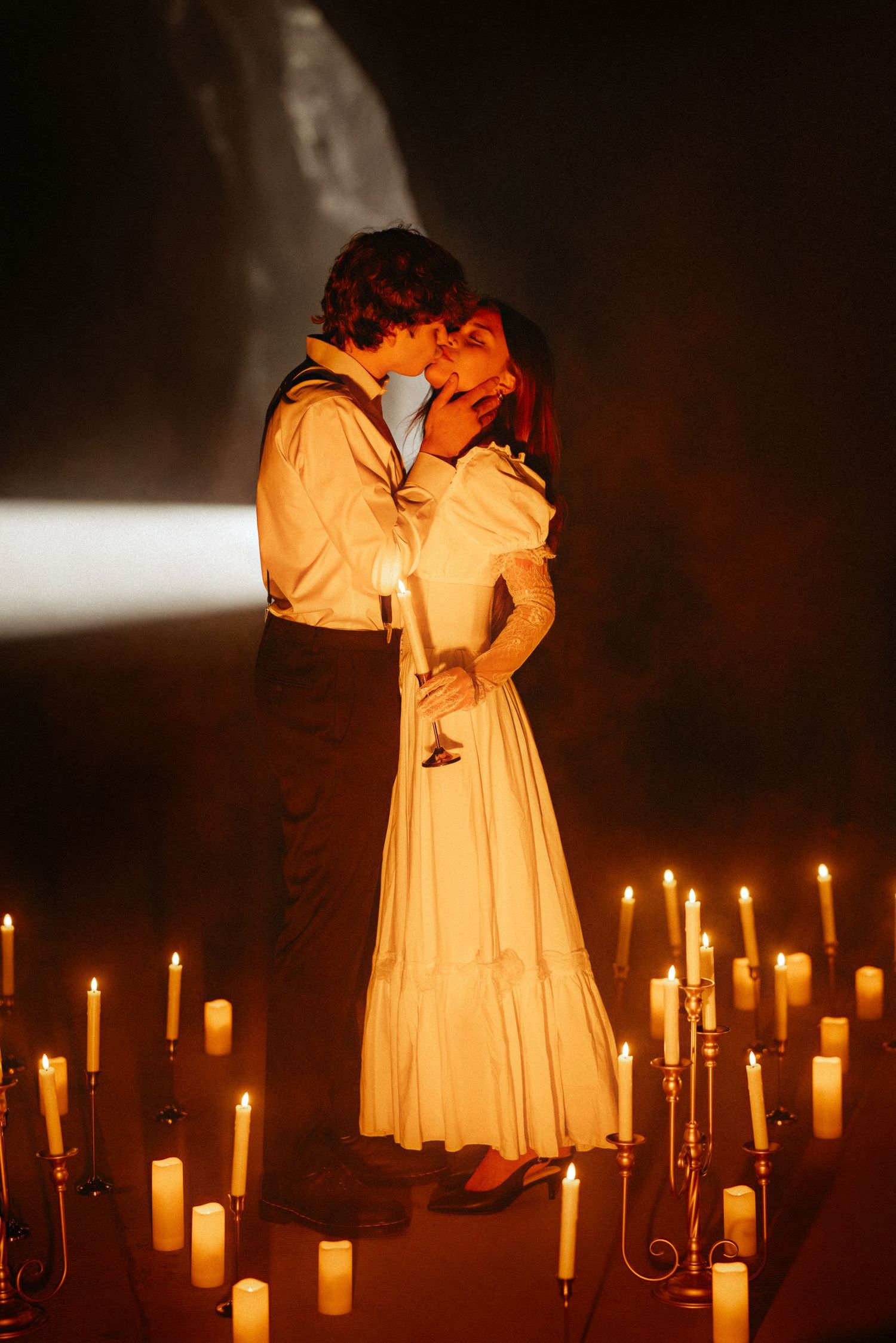 A row of glowing candles illuminates figures in white dresses standing in darkness creating an ethereal atmosphere.
