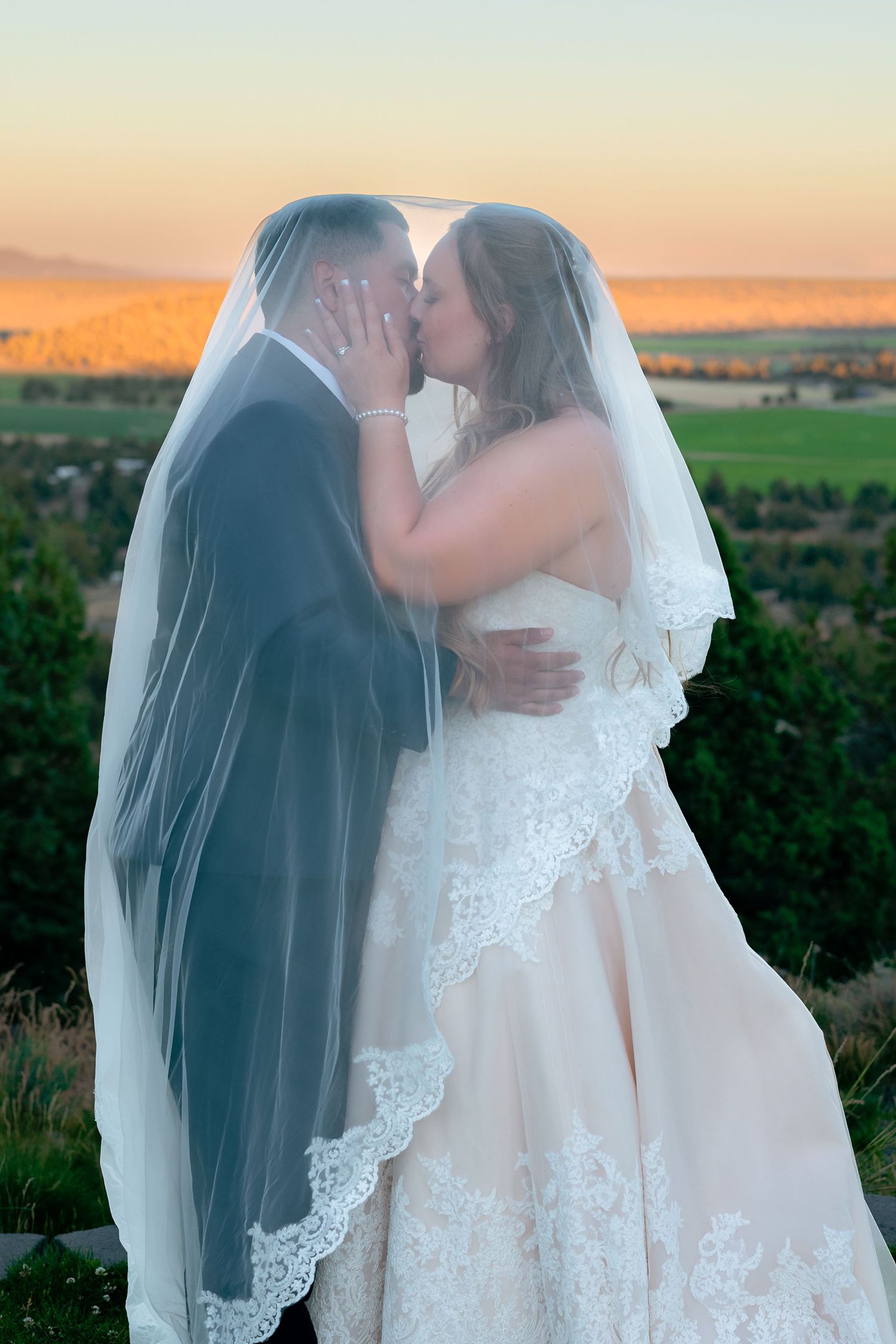 Bride and groom share a romantic kiss under a lace veil at sunset with scenic countryside views.