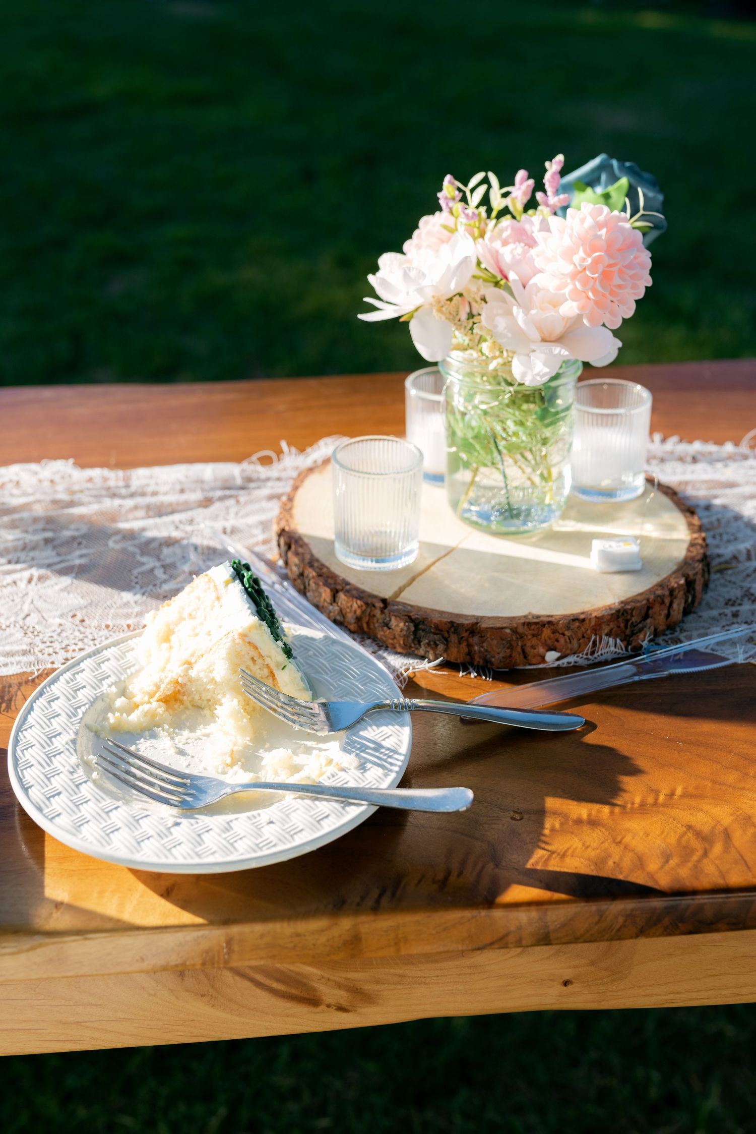 Slice of white wedding cake on a plate beside pink peonies on a rustic wooden table outdoors.