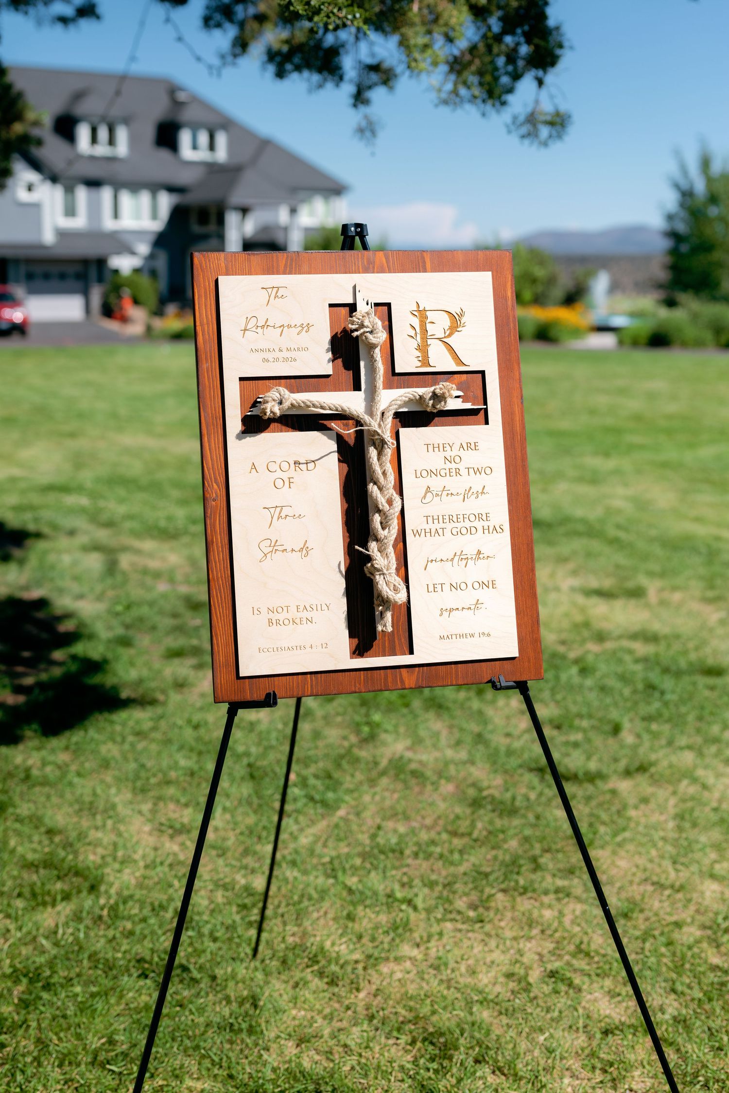 Wooden cross wedding sign with Bible verses and monogram displayed on easel outdoors.