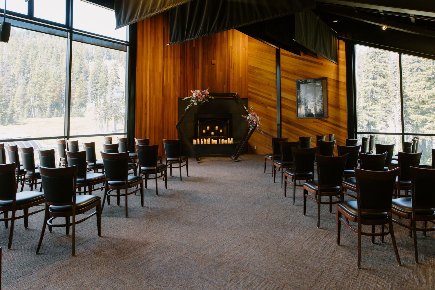 Elegant indoor wedding ceremony venue with wood-paneled walls, rows of dark chairs, and forest views through floor-to-ceiling windows.