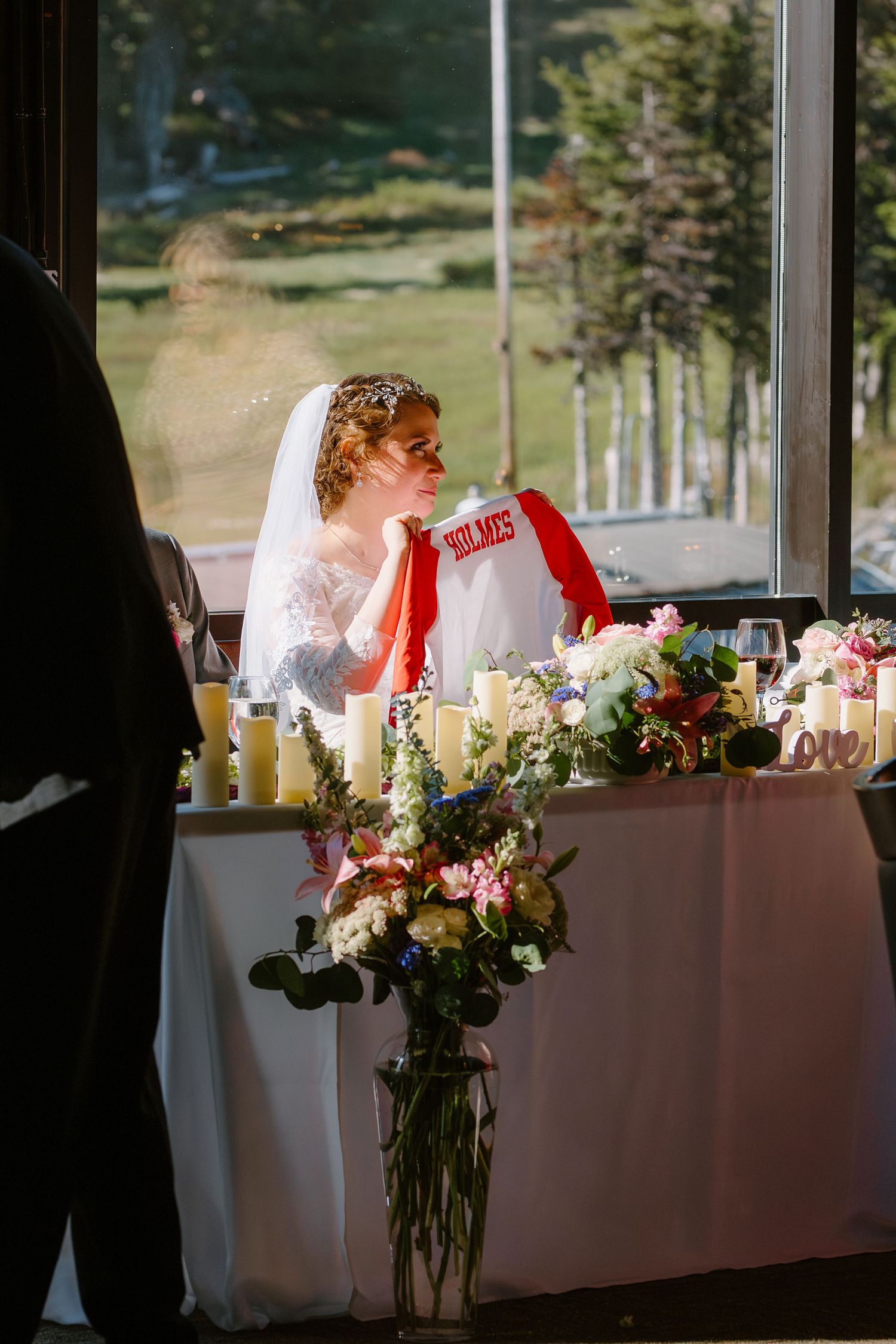 Bride in lace gown holds Holmes jersey at floral-decorated head table near large windows.