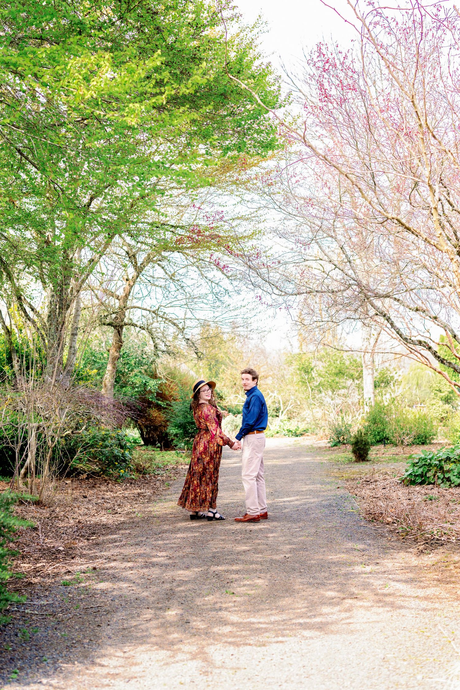 A couple walks along a cherry blossom-lined path in a spring garden setting.