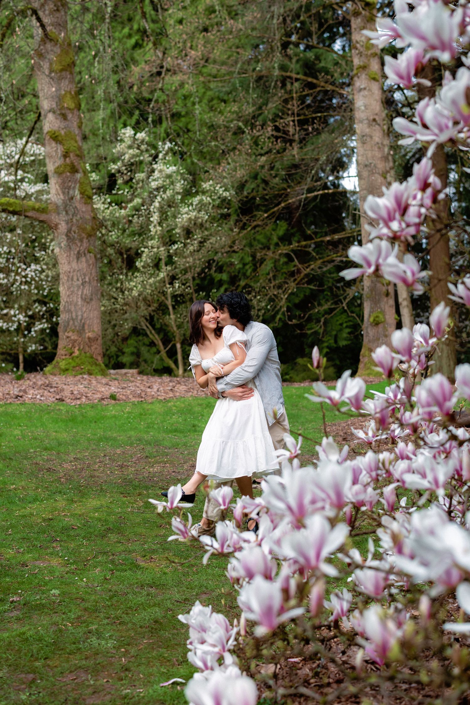 Two people share tender moments among blooming magnolia trees in a spring garden setting.
