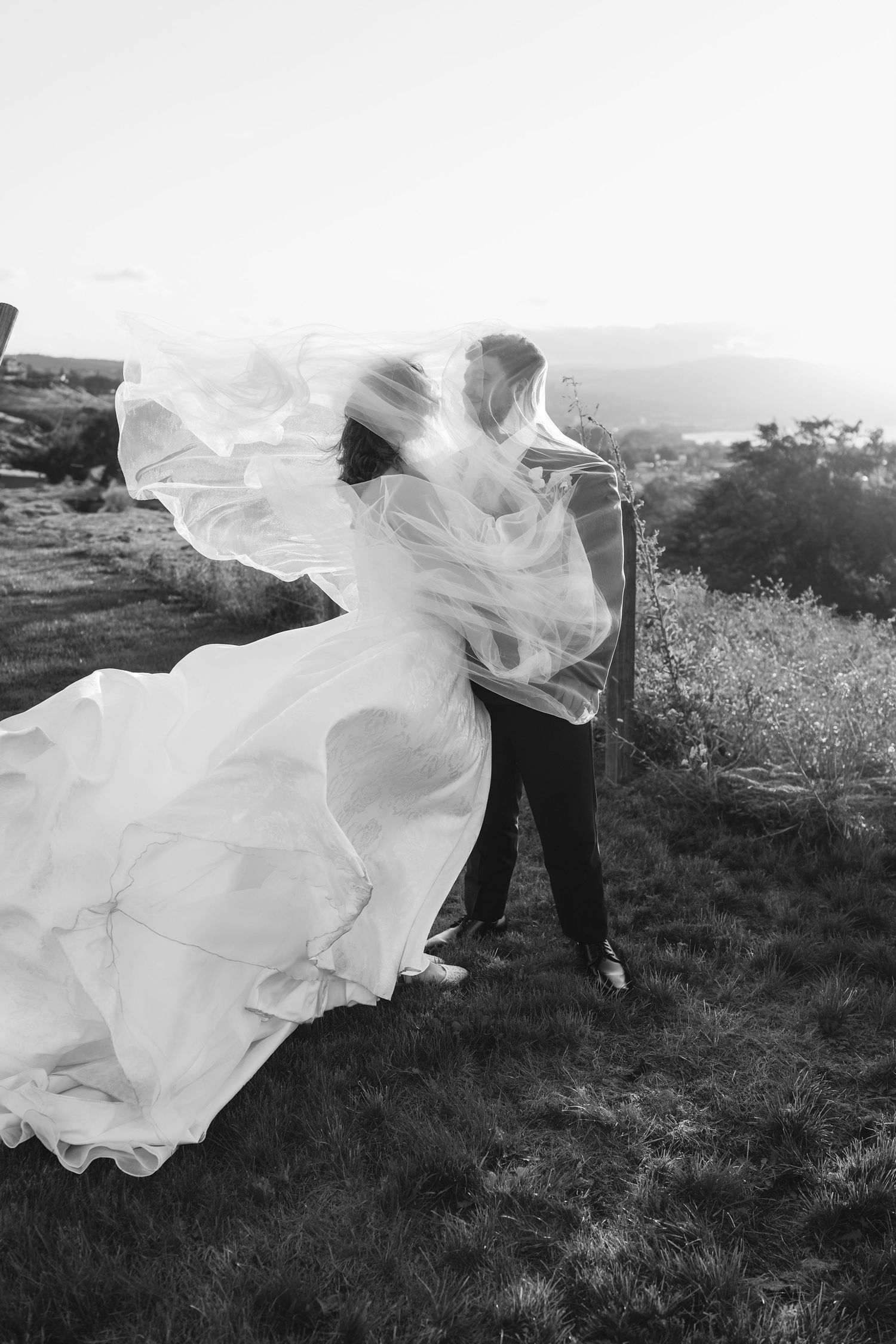 A couple in wedding attire dances outdoors as a white veil dramatically flows in the wind against a scenic landscape.