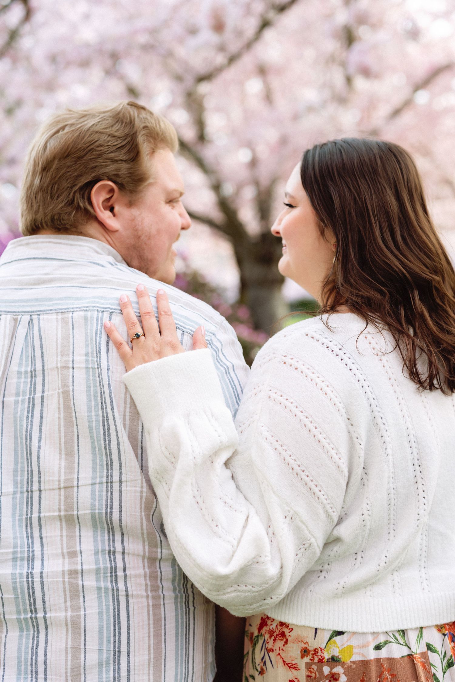 A couple shares tender moments in front of blooming cherry blossom trees wearing white sweaters.