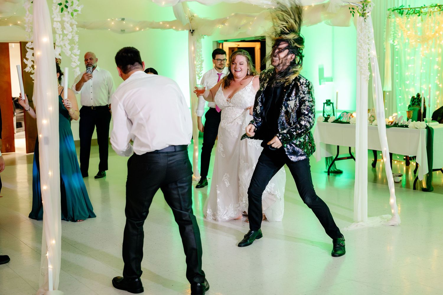 People dance and celebrate at a wedding reception with green uplighting illuminating the white venue interior.