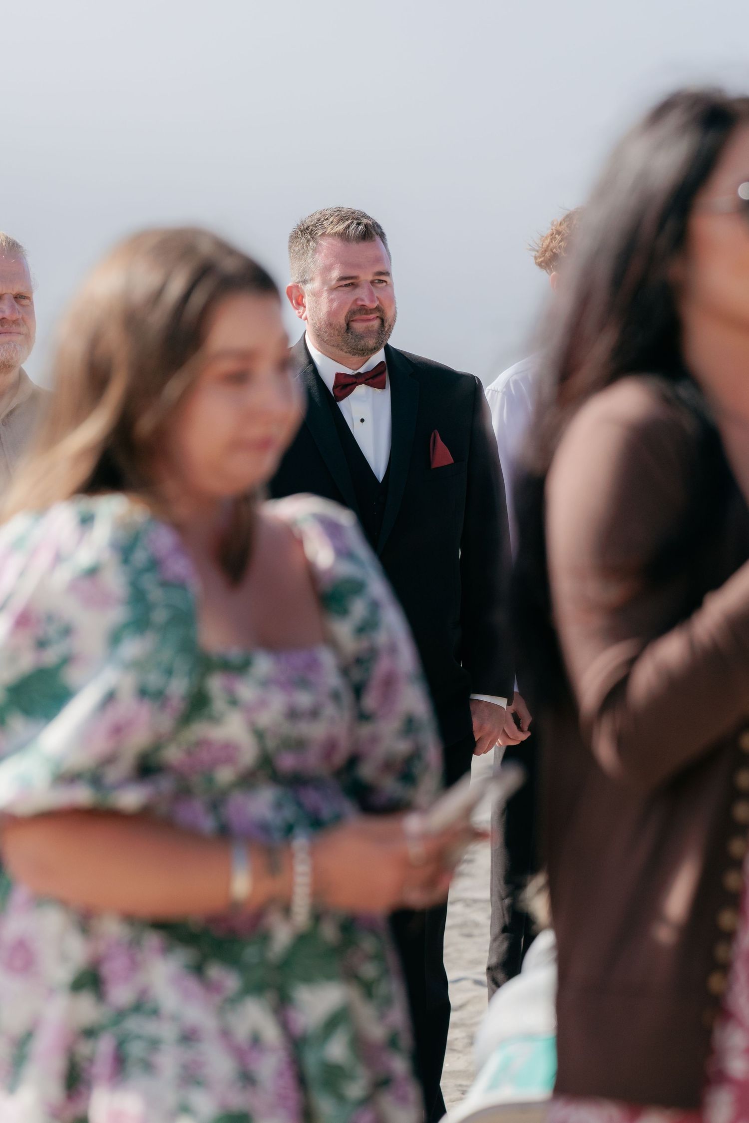 Wedding guests in foreground with formal attired attendee visible in background during outdoor ceremony.