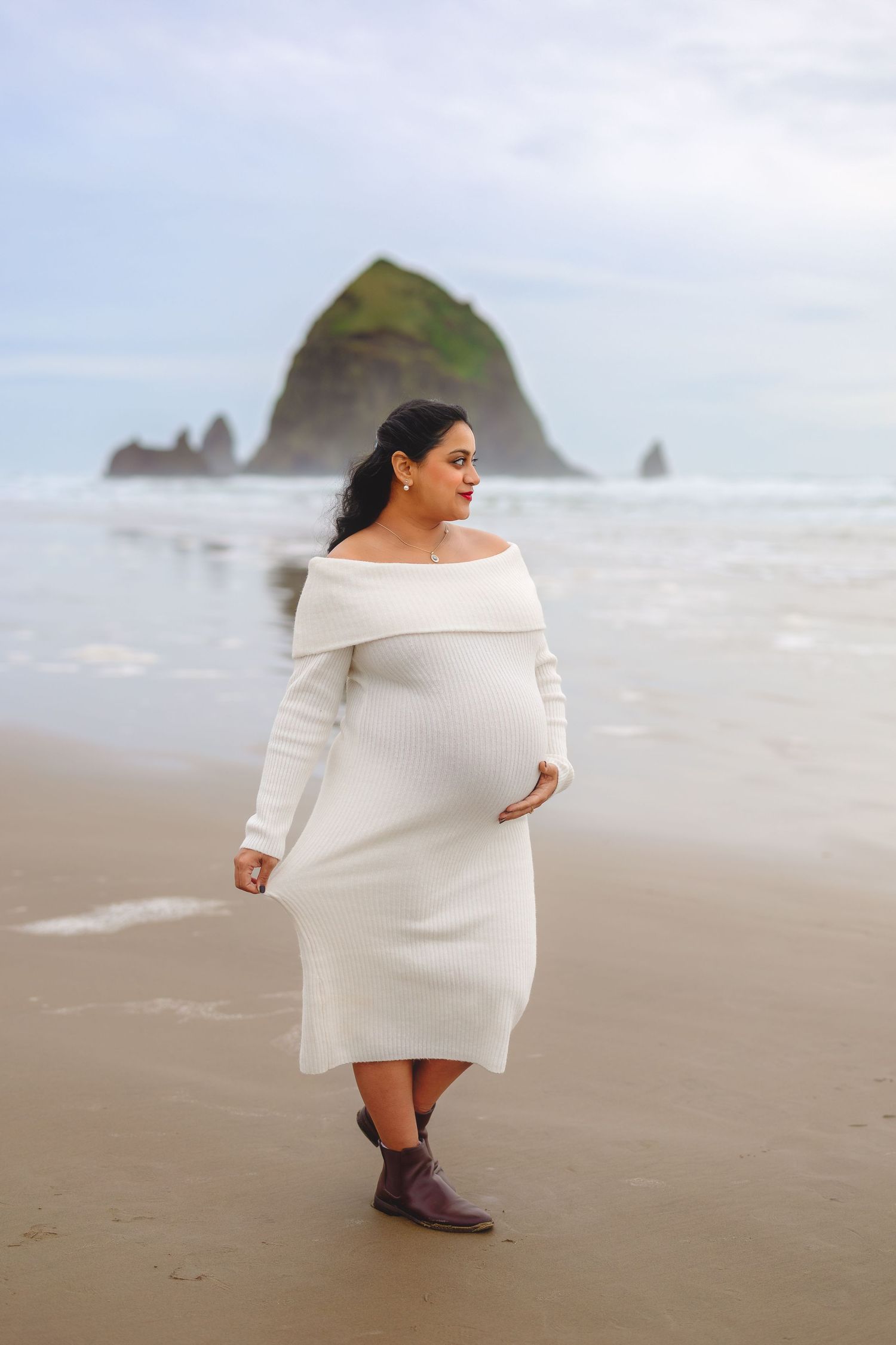 A figure in a white sweater dress poses on a misty beach with dramatic coastal rock formations in the background.
