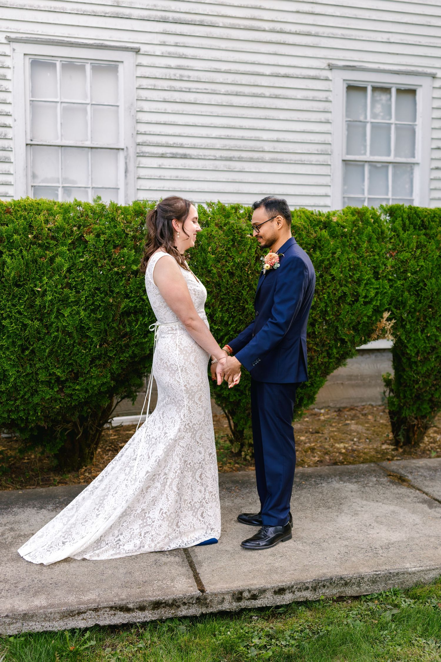 A couple in wedding attire dances playfully on a concrete path beside a white building and green hedge.