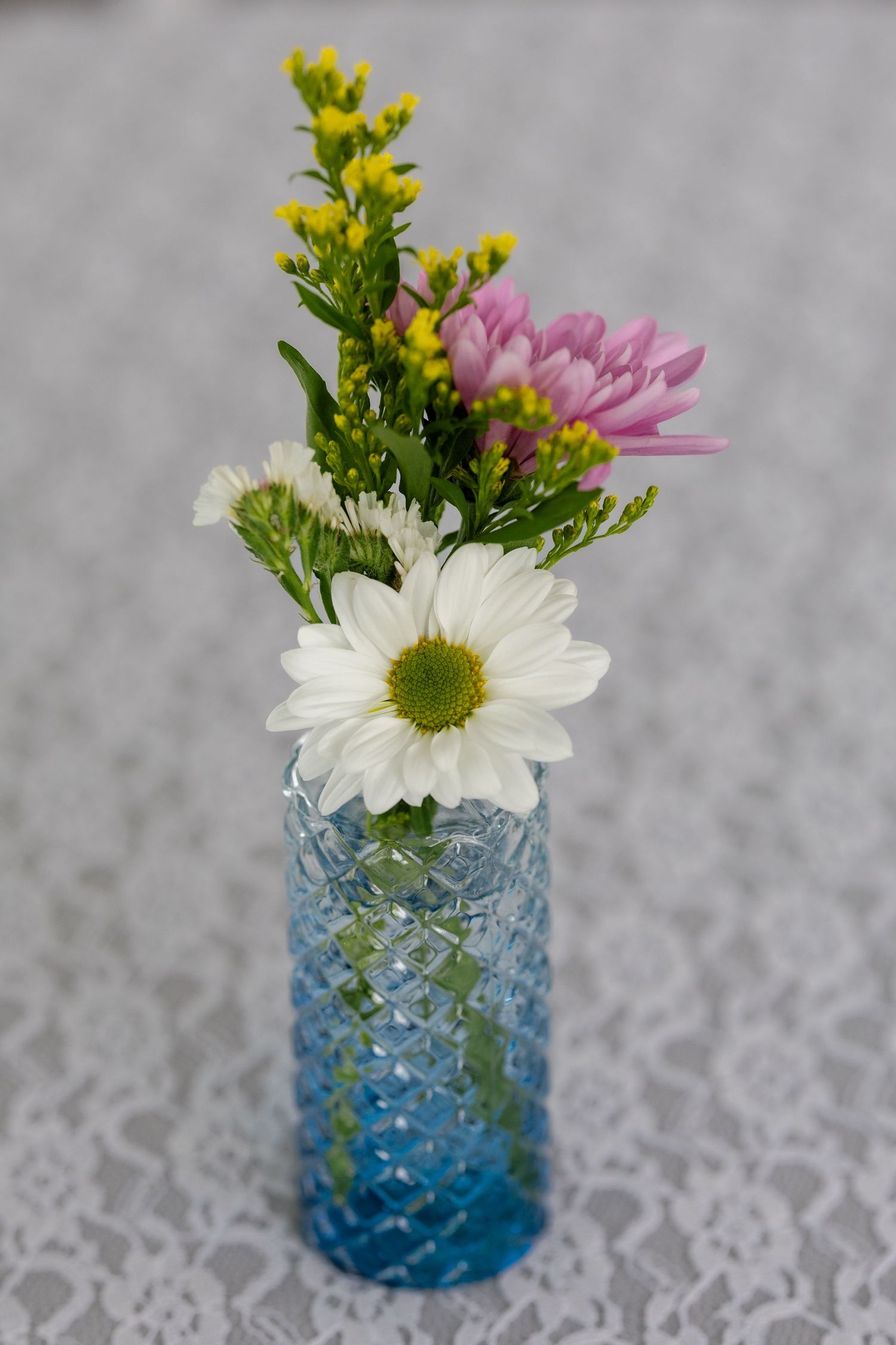 Small blue glass vase with white daisies and yellow flowers as wedding centerpiece decoration.