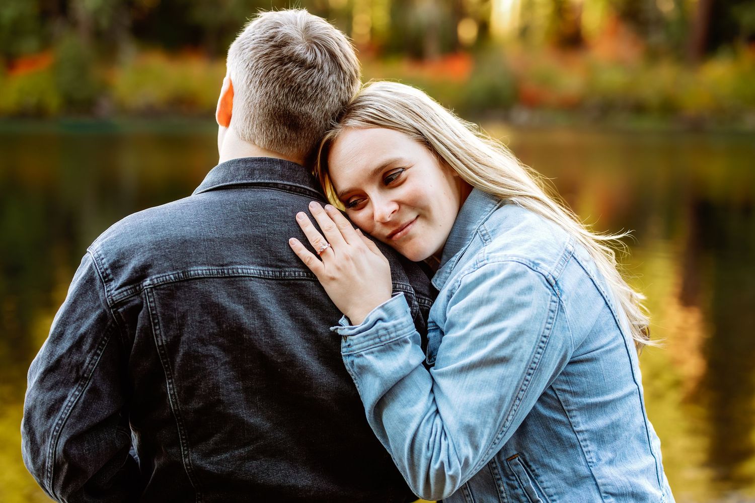 A couple embraces near a lake during golden hour with autumn trees reflecting in the calm water.