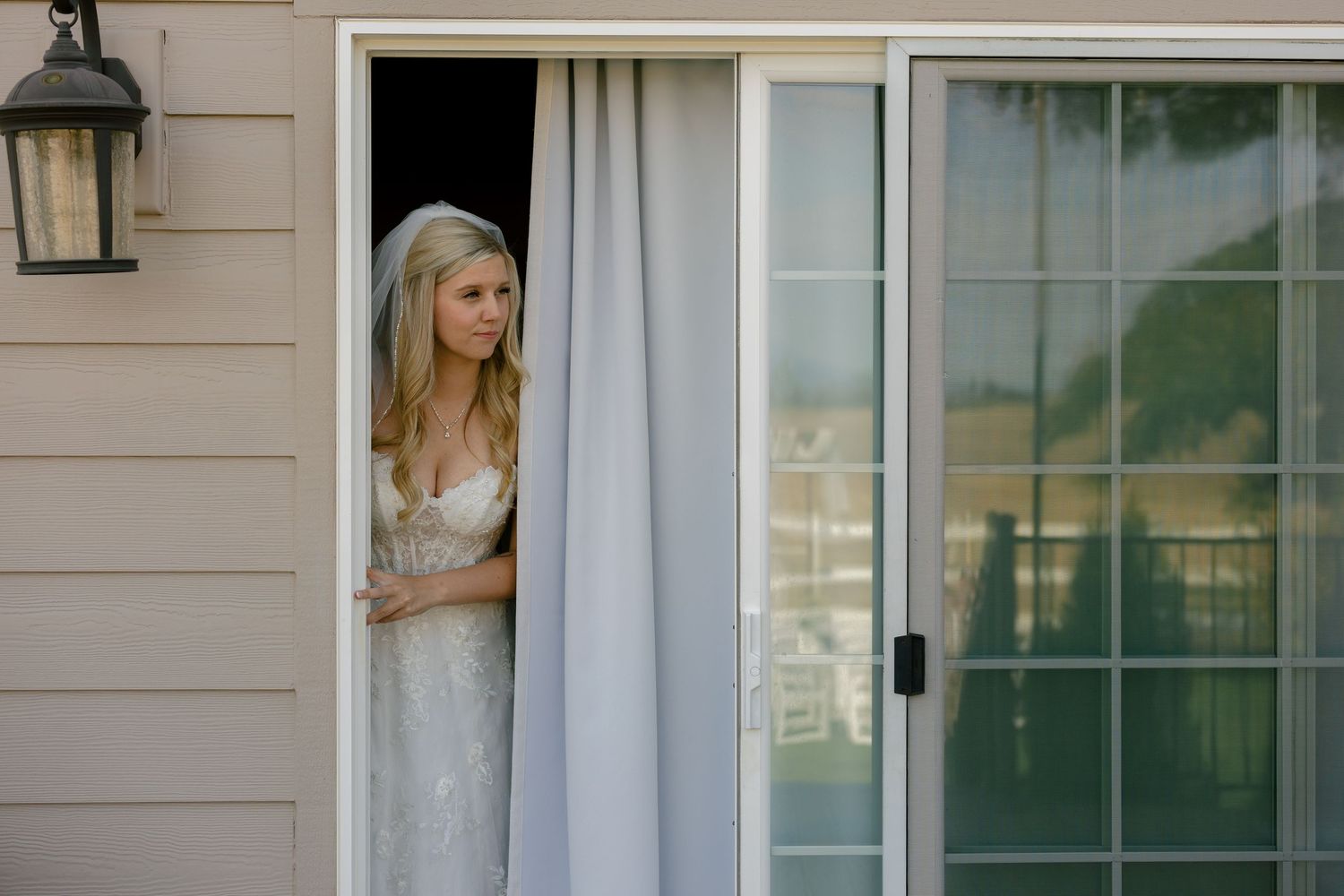 Bride in white dress looking through doorway with curtains and window.