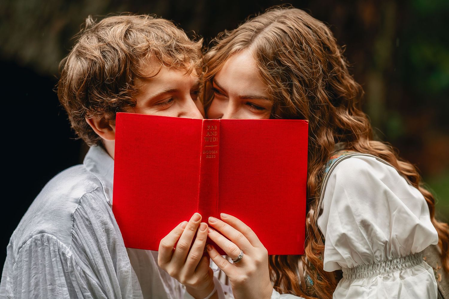 A couple shares a romantic moment while holding a red book in front of their faces outdoors.